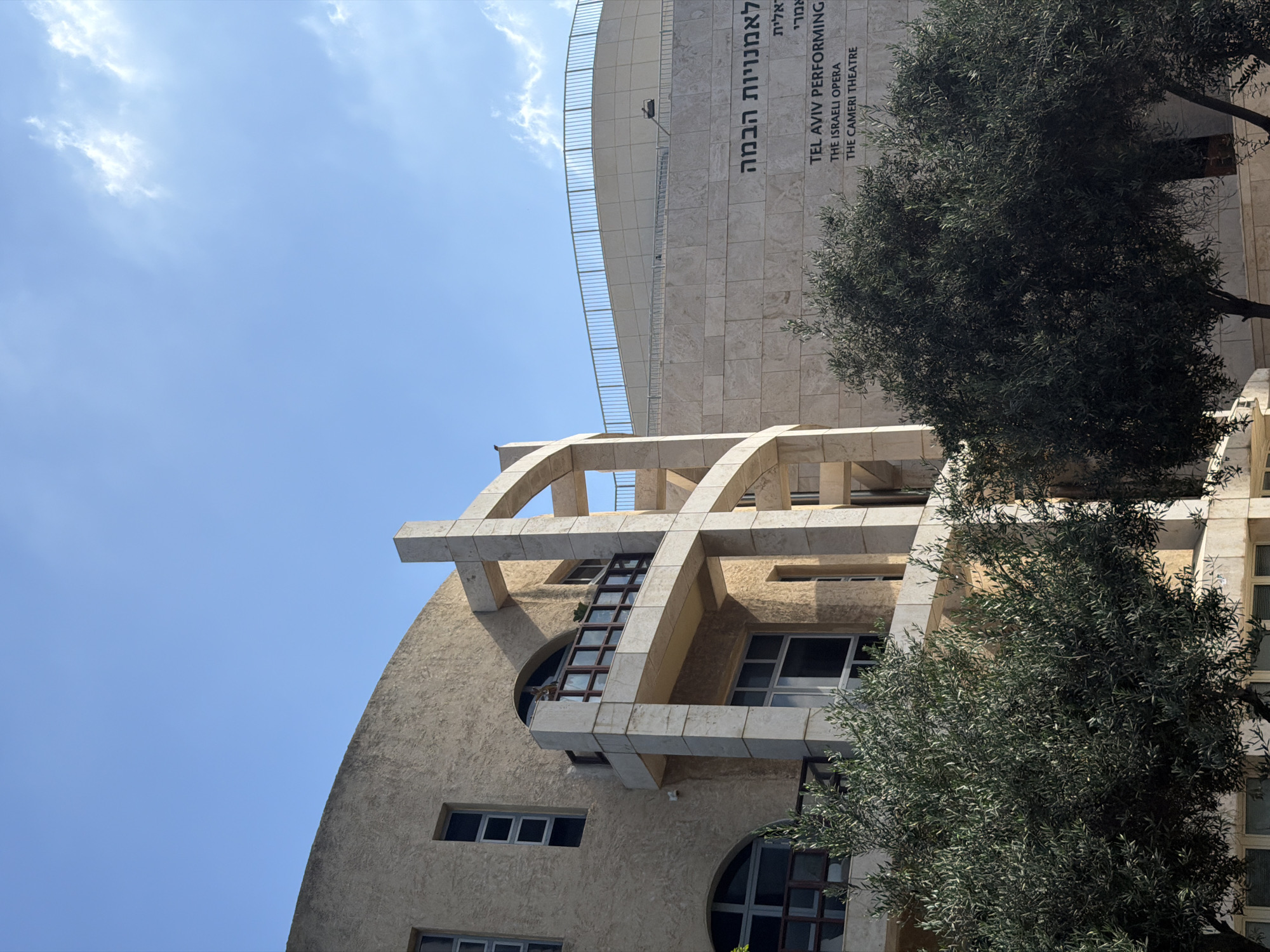 The image shows the exterior of the Tel Aviv Performing Arts Center, also known as the Israeli Opera and the Cameri Theatre, located in Tel Aviv, Israel. The building features a curved, modern architectural design with a light-colored stone facade. In the foreground, two olive trees with lush green foliage partially obscure the lower part of the building. The sky is a clear blue with a few wispy white clouds. The building's signage is visible on the right side, displaying "TEL AVIV PERFORMING THE ISRAELI OPERA THE CAMERI THEATRE" in English, and Hebrew text above it. Architectural elements such as balconies with wooden railings and large stone columns supporting arched structures are prominent on the left side of the building.