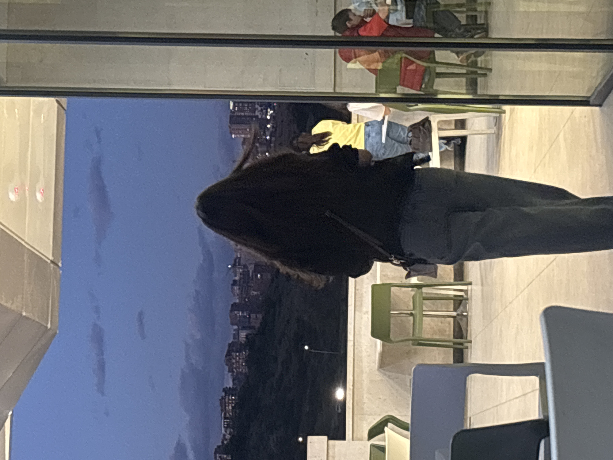 The image captures a twilight scene from what appears to be an elevated position in Yerevan, Armenia. In the foreground, a person with long dark hair, wearing a dark jacket and dark trousers, stands with their back to the camera, looking out towards the cityscape.
The scene is viewed through a glass partition or window, creating reflections of an interior space. In the upper part of the reflection, a person wearing a red top and light blue garment is visible, appearing to be lying down or leaning back, possibly looking at a phone. Below this, another person wearing a yellow top and blue jeans is reflected, seated. The reflections also show various chairs in shades of green, white, and blue, suggesting an indoor dining or lounge area.
Beyond the window, a dark blue sky dominates the upper portion of the exterior view, indicating dusk or early night. Below this, a dark, hilly landscape stretches out, dotted with scattered streetlights. Further in the distance, the city of Yerevan is visible, with numerous buildings illuminated by artificial lights, creating a sparkling horizon. The overall impression is of a quiet evening spent overlooking a city from a comfortable vantage point. No visible text is present in the image.