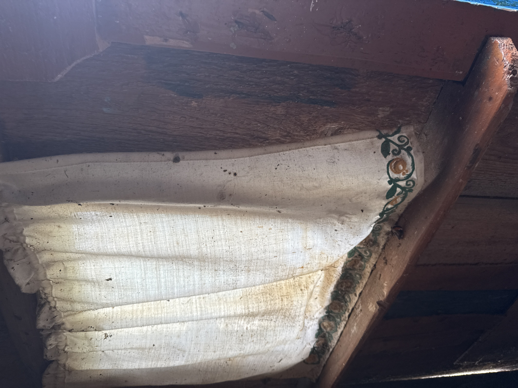 A white, dusty curtain with a floral trim hangs in front of a dark wooden wall. The curtain is gathered at the top, creating folds and pleats. Small dark specks, likely dust or debris, are visible on the fabric. The wooden wall behind the curtain has a rough texture and appears to be aged. The lighting suggests an indoor environment, possibly a window or a partition within a structure. The scene conveys a sense of disuse or neglect. There are no people or animals visible in the image.