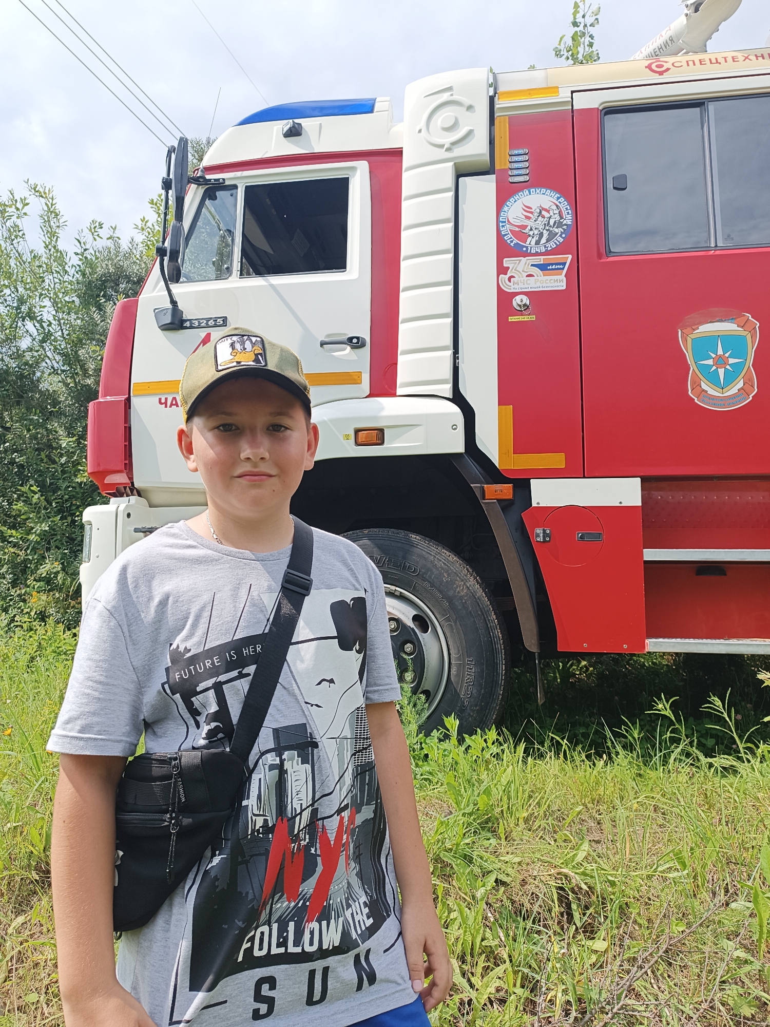 A young boy with light skin and short brown hair, wearing a grey t-shirt and a green baseball cap, stands in front of a red and white fire truck. He is looking directly at the camera with a neutral expression. The fire truck has various decals and markings on its side, including a circular emblem with figures and text, and a shield-shaped emblem. The boy is carrying a black crossbody bag. The scene is set outdoors, with lush green grass and trees in the background. The sky is overcast. The image is taken in the city of Chaikovsky, Russia.