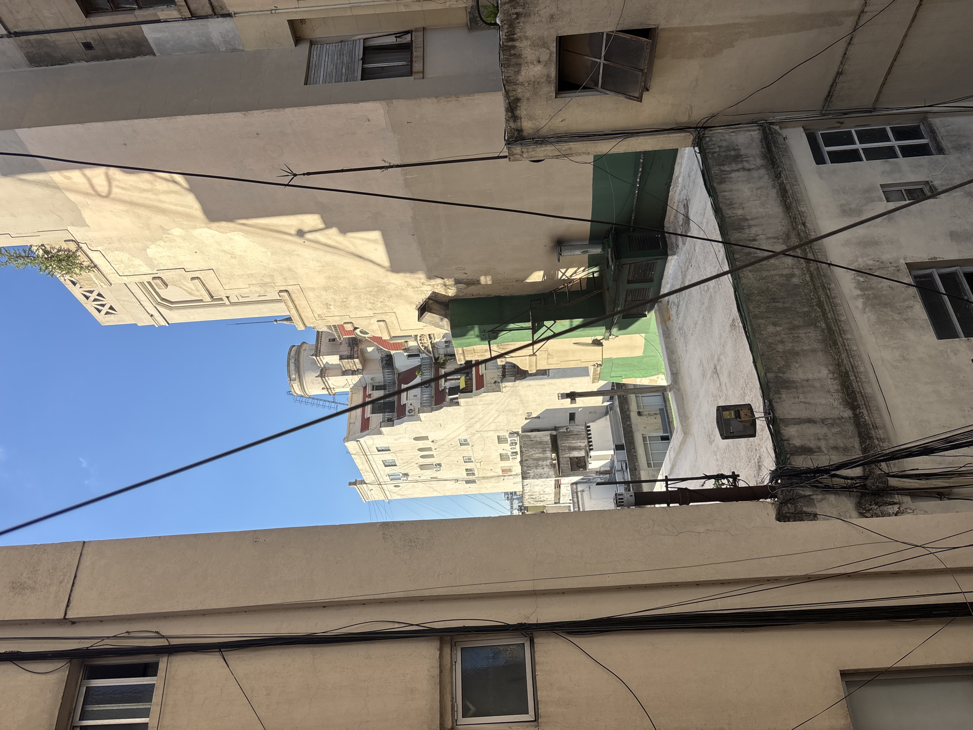 View of building exteriors in Buenos Aires, Argentina. Low-angle shot showing a narrow gap between buildings, revealing other structures and a patch of blue sky. Buildings are mostly beige and white, with visible windows, some open. A few buildings have green painted areas. Multiple overhead cables are strung across the space. The buildings appear aged, with some staining and weathered surfaces.