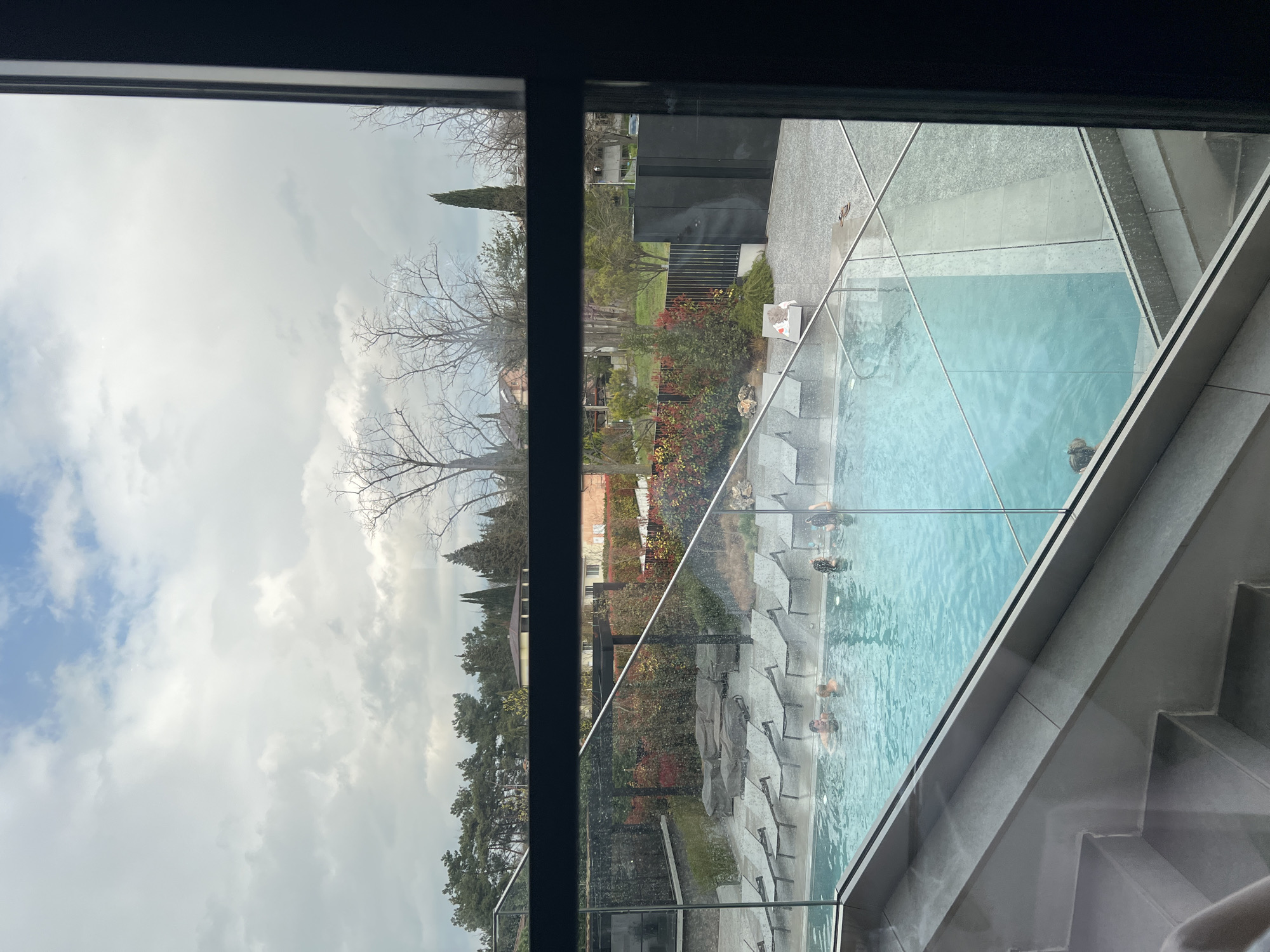 The view from a window shows a cloudy sky, trees, foliage, a building, and a pool with people in it. The pool is surrounded by lounge chairs and stonework. The location may be near Burgas, Bulgaria.