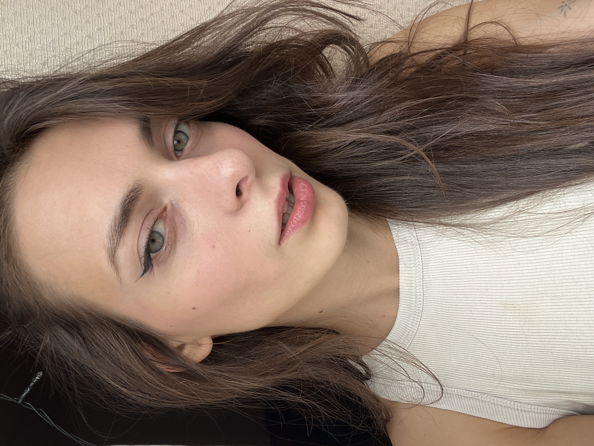 A woman with long brown hair, green eyes, and light skin is lying down, facing the camera. She is wearing a white ribbed tank top. The background appears to be a light-colored surface, possibly a carpet. There is no evidence to suggest the location is Krasnoyarsk, Russia.