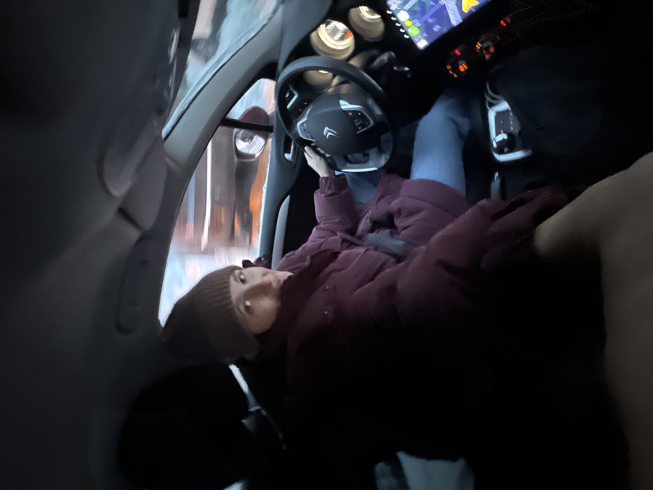 The image depicts the interior of a vehicle in Lesnoy Gorodok, Russia, with a female driver in motion, looking back at the camera. She is wearing a brown knit beanie and a dark maroon puffy winter jacket over light blue pants. Her right hand is positioned on the black, multi-function steering wheel, which features a Citroën logo. To the right of the steering wheel, circular instrument gauges are visible, and below them, an illuminated infotainment screen displays a navigation map interface with green, yellow, and red lines. The exterior background, visible through the windshield, is horizontally blurred, indicating vehicle movement. Snow is present on the ground outside, suggesting winter conditions and low ambient light. A hand, belonging to the front passenger (presumed photographer), is visible in the lower left foreground.