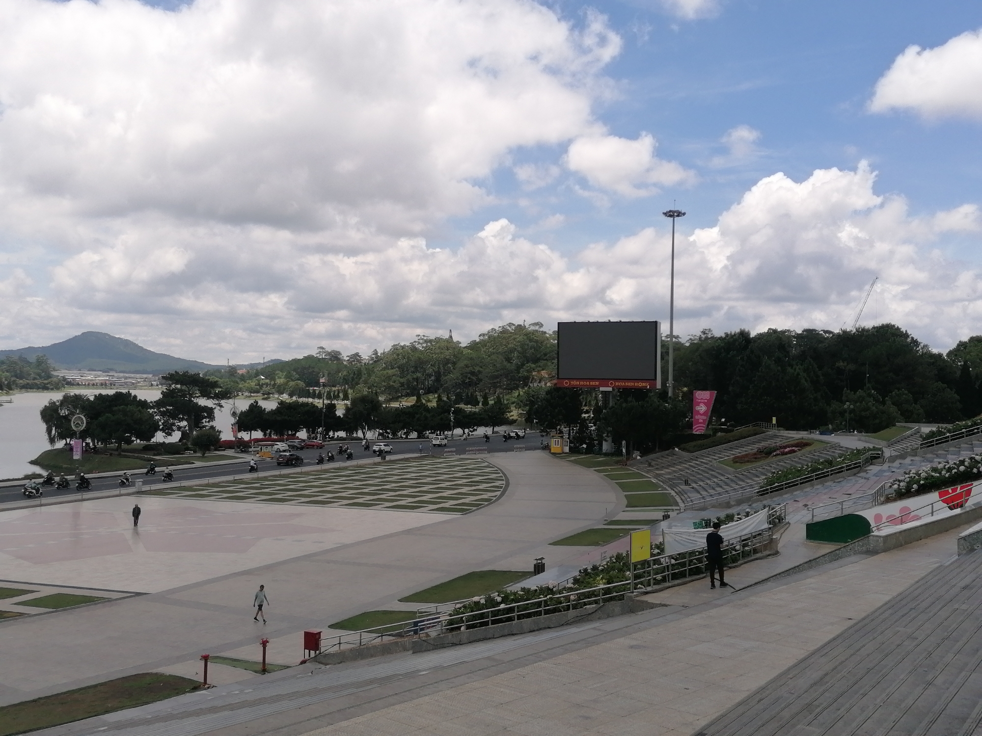 The image depicts a view of Lam Vien Square in Da Lat, Vietnam, characterized by a large open public space and its surroundings. The sky is cloudy, casting soft light over the scene. The foreground shows a concrete stairway leading down to the square. A metal railing lines the edge.
A person wearing dark clothing stands near the railing, and appears to be holding a long object such as a broom. There is another man walking alone in the square, and one more distant man walking near the lake. In the middle distance, the large open square is evident, with a lighter grey patterned design on its surface. Behind the square, there is a road with traffic, including cars and motorcycles. Beyond the road is a lake, and trees along the shore. A hill or small mountain can be seen in the distance. To the right of the square is tiered seating and planting beds.
Dominating the skyline is a tall light post, and a large LED screen with advertisements in Vietnamese. Green trees fill the scene. There is also a construction crane visible to the right of the screen.