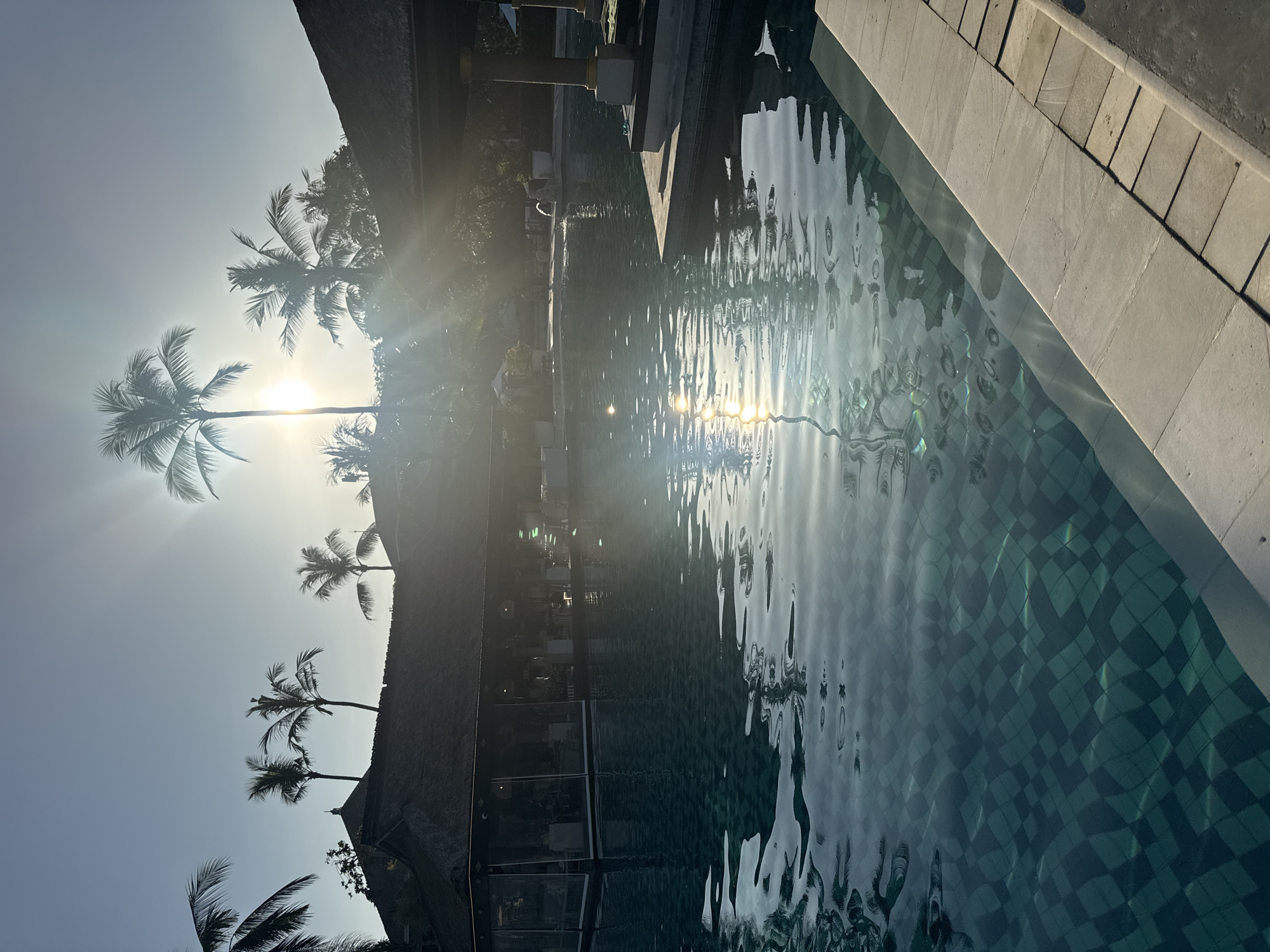 The image, taken in Kuta Selatan, Indonesia, shows a sunlit swimming pool reflecting a building facade and palm trees. The pool water is tiled in a checkered pattern, and sunlight reflects off its surface. The building facade is silhouetted, and several palm trees are visible against a bright sky. The sun's glare dominates the sky, positioned behind one of the palm trees. A stone border edges the pool on the right side.
