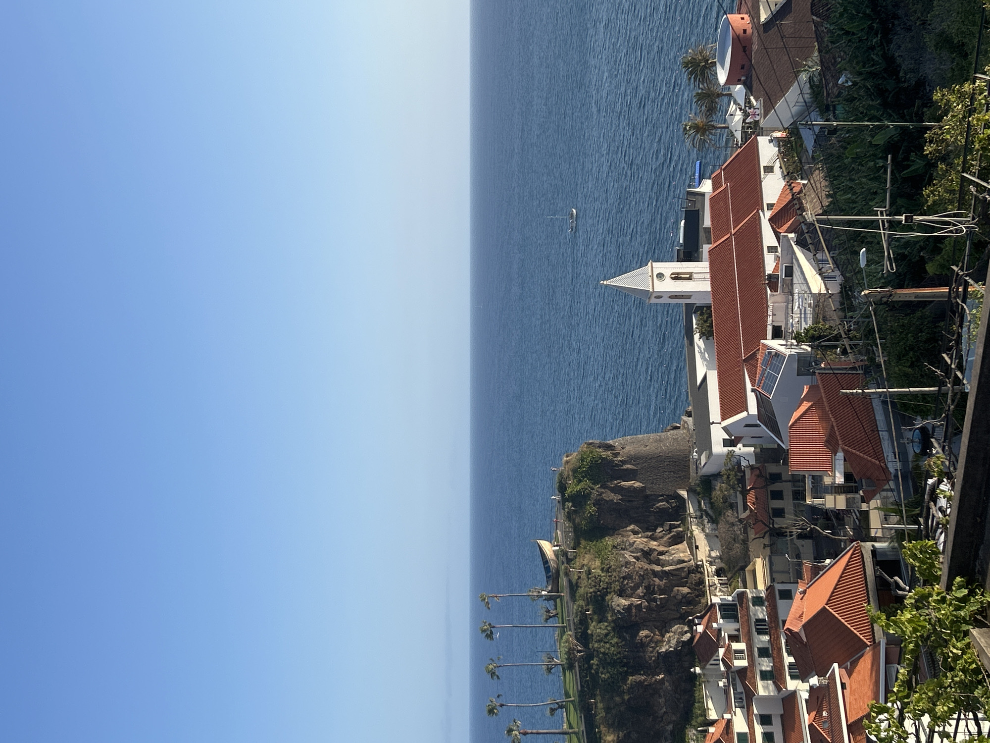 The image shows a coastal view from Câmara de Lobos, Portugal. The scene includes a blue sky and ocean, with a small boat visible on the water. Along the coast, there are palm trees, cliffs, and buildings with red-tiled roofs. A church with a white tower is also visible. The buildings are situated on the cliffside and extend towards the right edge of the image.