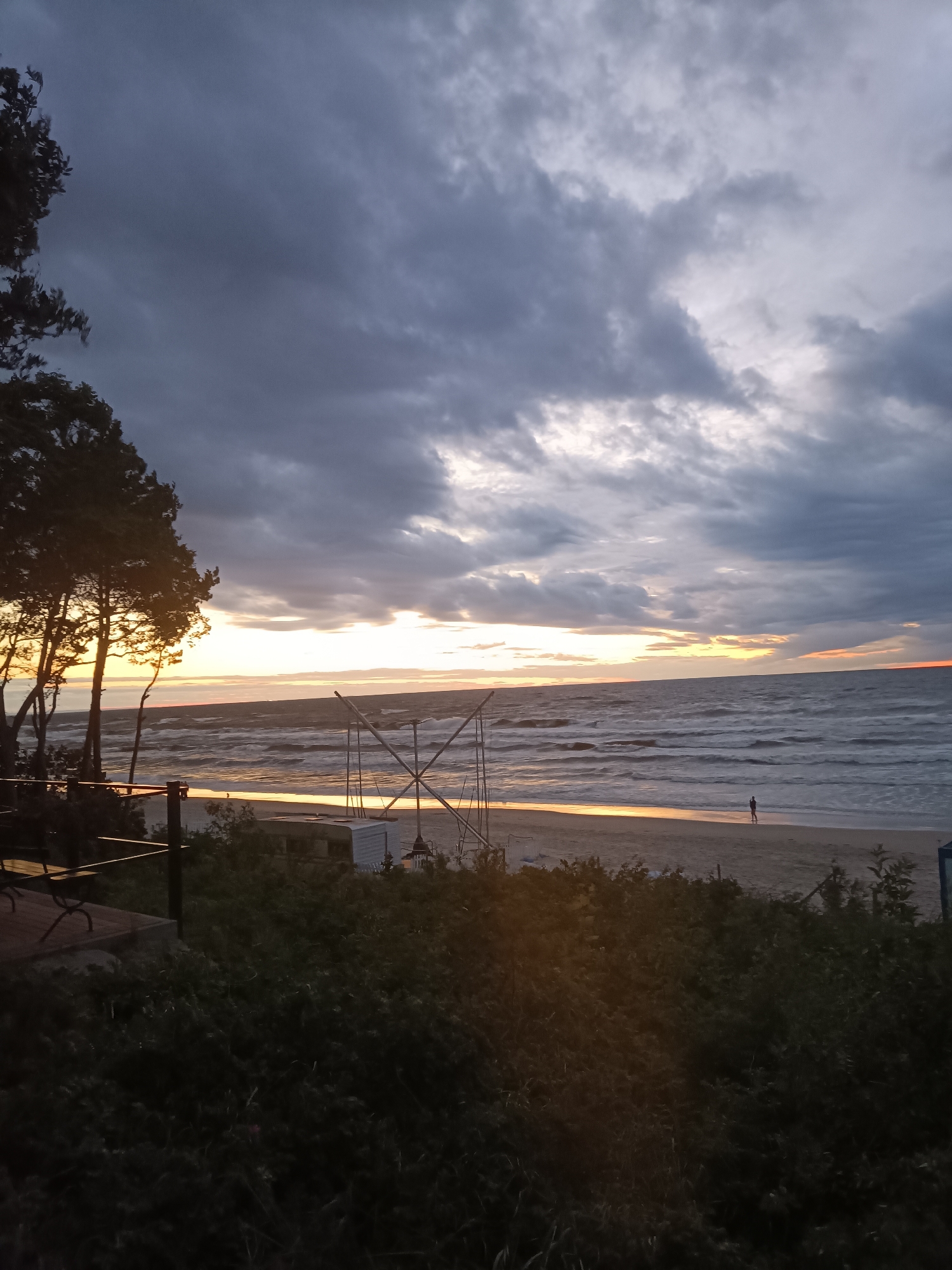 The image depicts a scene at Stegna beach in Poland during a cloudy sunset. In the foreground, dense green bushes partially obscure a wooden deck with benches and railings. Beyond the vegetation, a sandy beach stretches towards the sea, where waves are breaking. A lone figure, silhouetted and small, is walking on the wet sand near the water's edge. Further down the beach, a white trailer or small building is visible, along with some metal structures that might be part of an amusement or playground. The sky is filled with heavy, dark clouds, but a bright band of orange and yellow light emanates from the horizon where the sun is setting. Pine trees are present on the left side of the frame, with their silhouettes backlit by the sunset. The overall atmosphere is moody due to the cloudy sky and the approaching dusk.