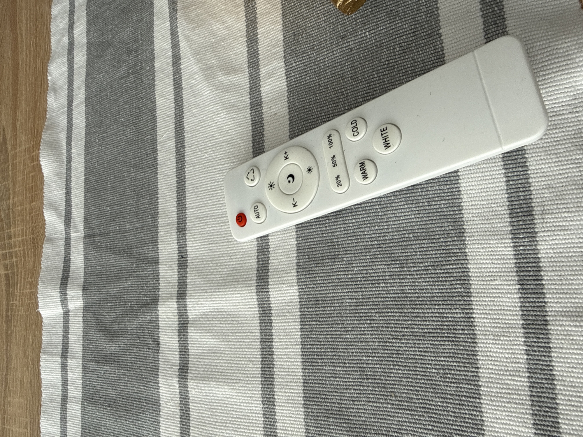 A close-up shot presents a white remote control lying on a horizontally striped grey and white textile surface. A small section of light-colored wood flooring is visible in the lower-left corner. The remote features several buttons including "AUTO", "K-", "K+", "20% 50% 100%", "WARM", "COLD", and "WHITE". The scene appears indoors under ambient lighting.