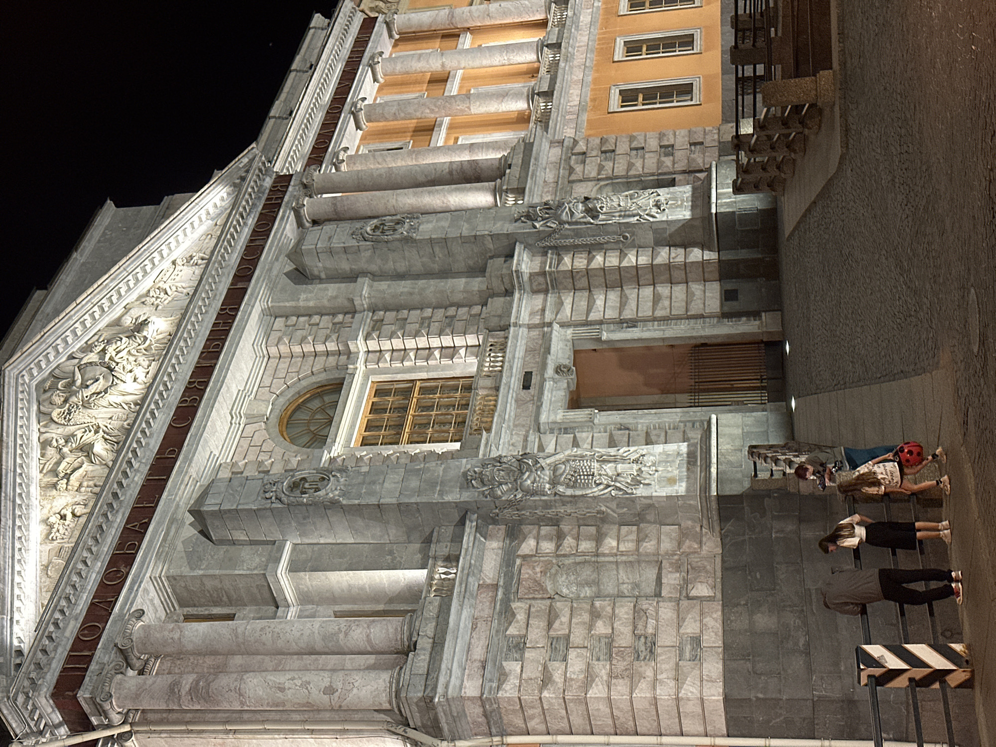 The image captures a nighttime view of the Singer House, also known as the Dom Knigi, located on Nevsky Prospekt in Saint Petersburg, Russia. The iconic building, illuminated by spotlights, showcases its Baroque Revival architecture with ornate stone carvings and a distinctive glass globe atop its dome. The foreground features a cobblestone street and a group of four individuals standing near a railing. Three of them appear to be young women and one appears to be a young man. One of the young women is wearing a white t-shirt and a black skirt, another is wearing a patterned dress, and the third is wearing a light-colored t-shirt and dark pants. The young man is wearing a grey hoodie and dark pants. They are looking towards the building or engaging in conversation. The overall scene conveys a sense of evening ambiance in a historical urban setting.