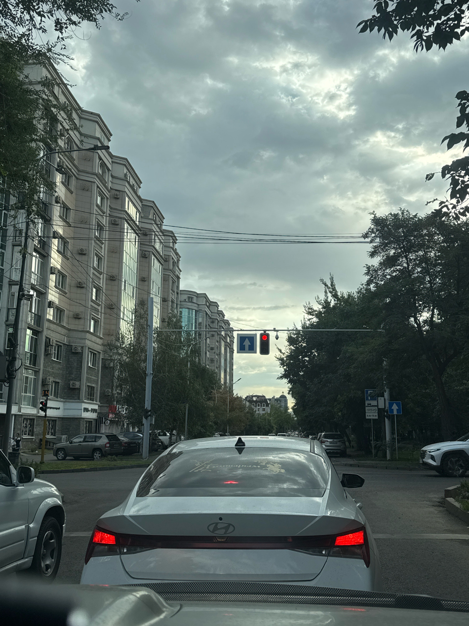 The image captures a view from a vehicle in traffic on a street in Almaty, Kazakhstan. A row of modern, light-colored apartment buildings with numerous windows and balconies lines the left side of the street. Trees and foliage are abundant on the right side, with dense green canopies and some taller trees. A traffic light hangs above the intersection, displaying a red light for straight-ahead traffic, indicated by an illuminated red circle and a blue arrow pointing upwards. Several cars are stopped at the red light, including the vehicle from which the photo is taken, which is following a white Hyundai sedan. Other vehicles, including a silver SUV, are visible in the distance and on the right side of the road. Overhead power lines crisscross the sky, and the sky itself is overcast with dramatic clouds. Street signs are visible, including one with a parking symbol and hours of operation.