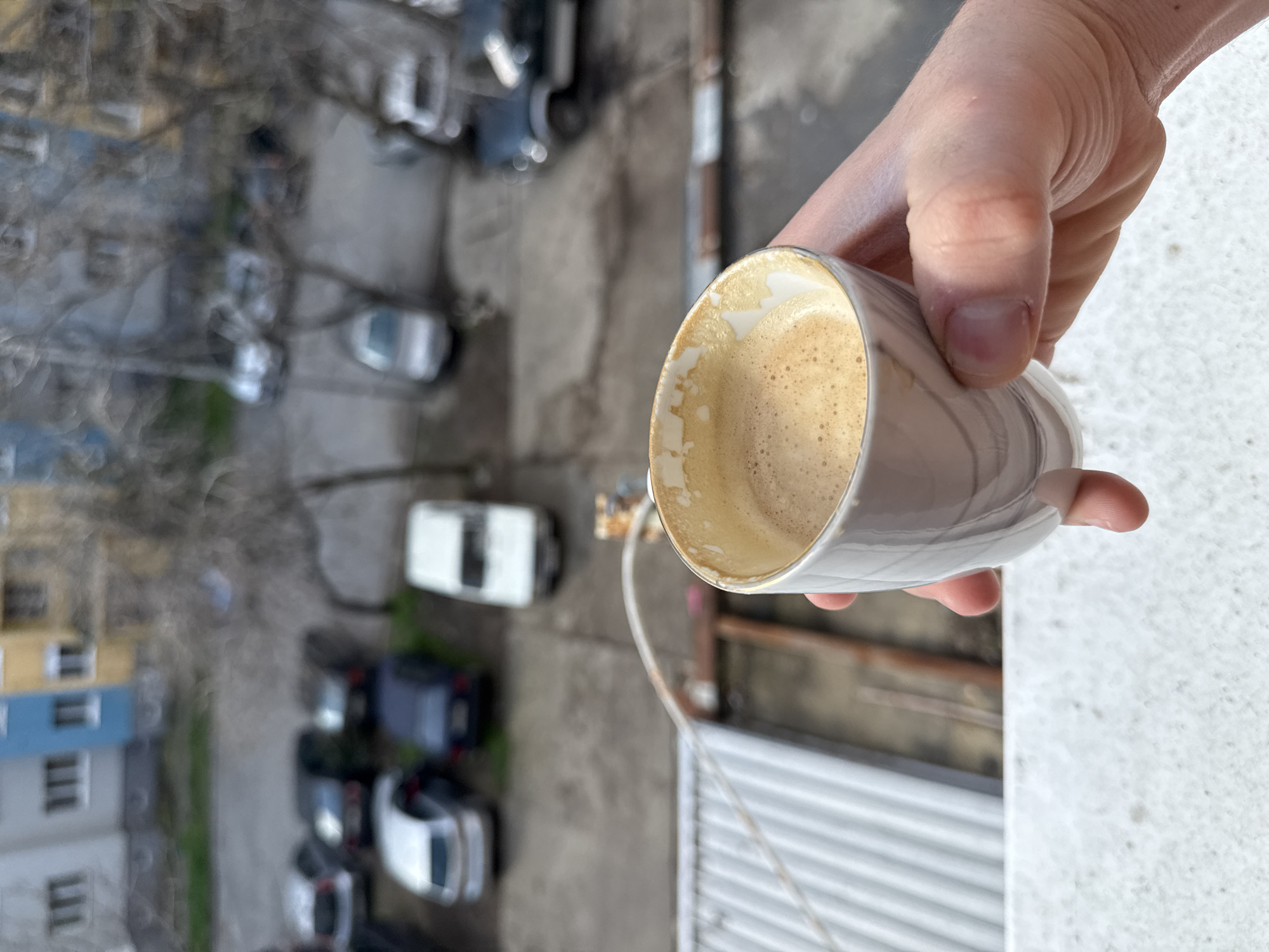 A hand holds a cup of coffee over a balcony. Below is a view of a parking lot filled with cars, some bare trees, and a building with blue and white trim. The location is plausibly Stara Zagora, Bulgaria, based on common architectural styles.