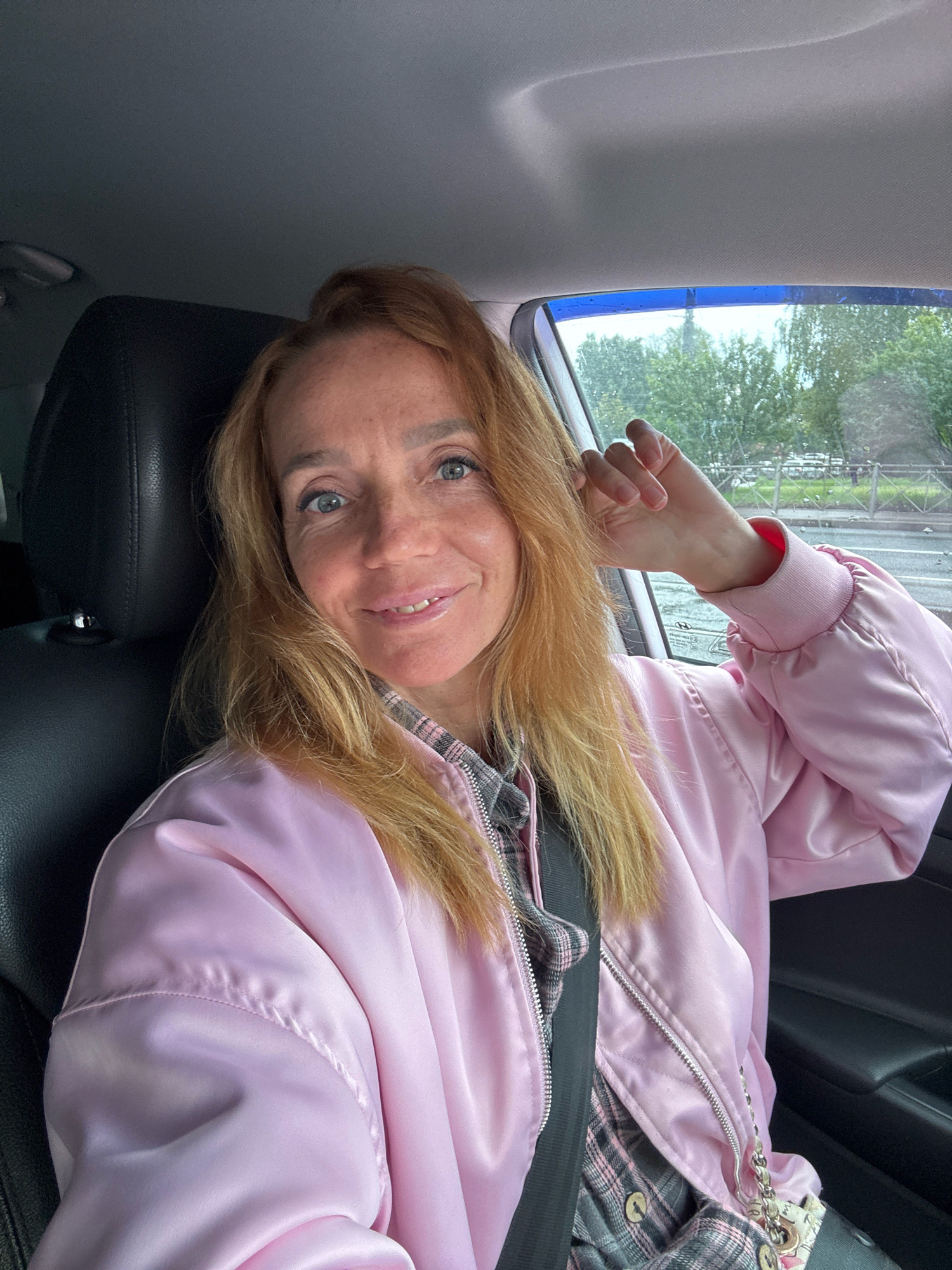 A woman with shoulder-length blonde hair, wearing a pink satin bomber jacket and a plaid shirt, is seated in the driver's seat of a car. She is looking at the camera and smiling, with her right arm resting on the window frame. The interior of the car is visible, including a black leather seat and a portion of the dashboard. Through the window, trees and a fence can be seen, suggesting an outdoor environment. The overall lighting indicates daylight. The scene appears to be in Saint Petersburg, Russia, given the context of the request.
