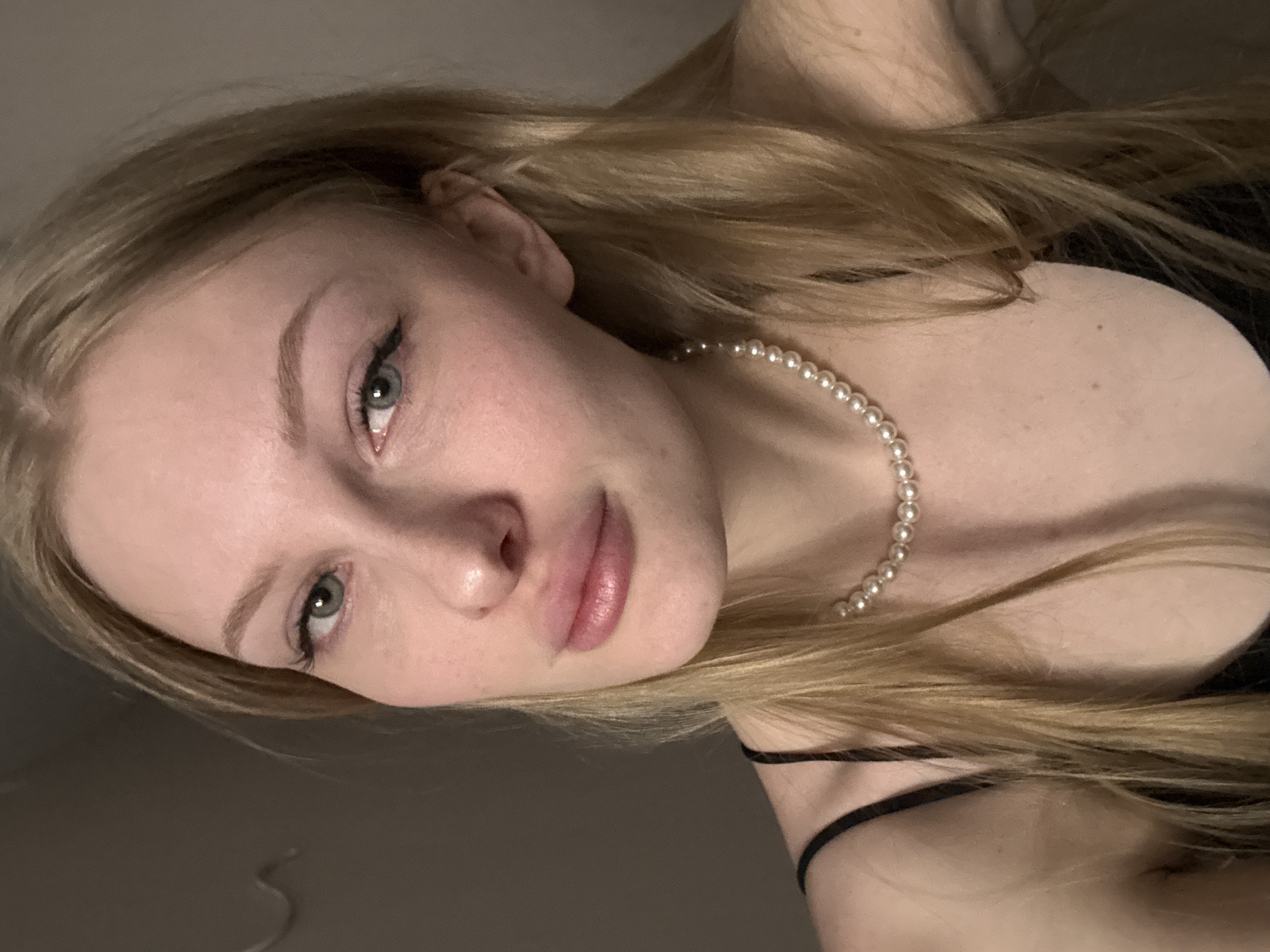 The image is a close-up, head-and-shoulders shot of a young Caucasian woman with blonde hair, pale skin, and light blue eyes. She is wearing a black spaghetti-strap top and a pearl necklace. Her lips are full and painted with a shimmery pink lipstick. Her eyebrows are well-defined and arched, and she has a subtle winged eyeliner. The background is a plain, neutral-toned wall, which is out of focus. There is no indication of the city of Perm, Russia, in the image.
