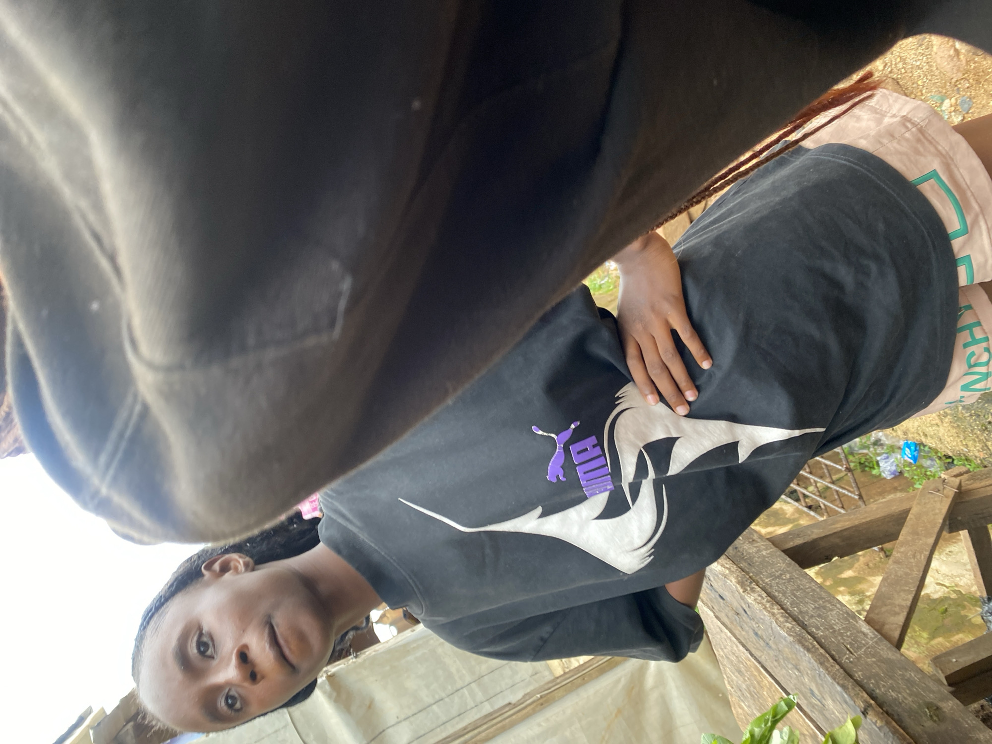 A low-angle, close-up shot captures a young Black individual standing outdoors, possibly in a peri-urban or rural setting within Jos, Nigeria. The subject is wearing a black t-shirt with a prominent white and purple Puma logo and light-colored shorts with a green trim and text. Their right hand is resting on their hip, and their left hand is not visible. The background consists of wooden structures, possibly a makeshift stall or fence, with some greenery and what appears to be a dirt ground. The overall lighting suggests an overcast day. The camera appears to be held by another person, partially visible in the foreground, obscuring the lower right portion of the frame with their dark clothing.