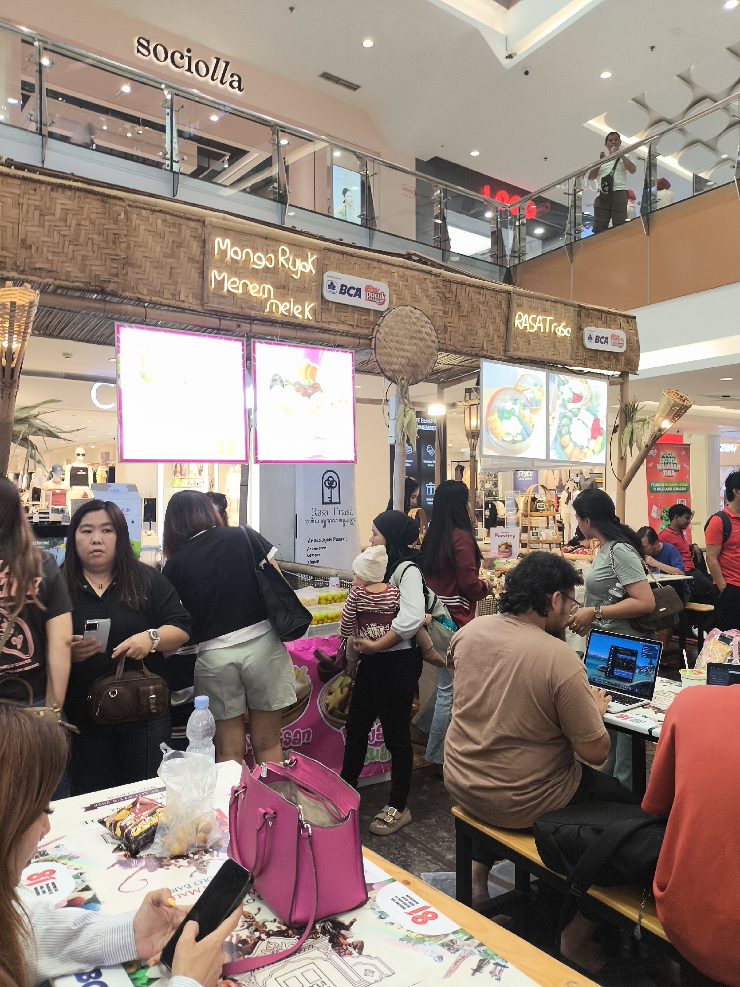 The image depicts a bustling indoor market or food court likely located in Grogol, Indonesia, possibly at a mall like Central Park Mall. In the foreground, a woman with long brown hair is seated at a table, engrossed in her smartphone. On the table, there's a bright pink handbag, a plastic bag of snacks, and a bottle of water. To her right, another person in an orange shirt is seated, looking away from the camera.
Behind them, a man with dark curly hair is seated at a table, working on a laptop. Several other individuals are standing and milling about, some appearing to be shoppers or vendors. Above the crowd, a thatched-like structure with neon signs is visible. One sign reads "sociolla" in a stylized font, suggesting a beauty or lifestyle store. Another sign features neon text in what appears to be Indonesian: "Mango Ryok Merem Melek," and "RASAT rasa." Two large illuminated displays showcase colorful food items. The environment is brightly lit, typical of an indoor commercial space, with a ceiling visible in the upper portion of the frame. The overall scene suggests a lively commercial atmosphere with food and shopping activities.