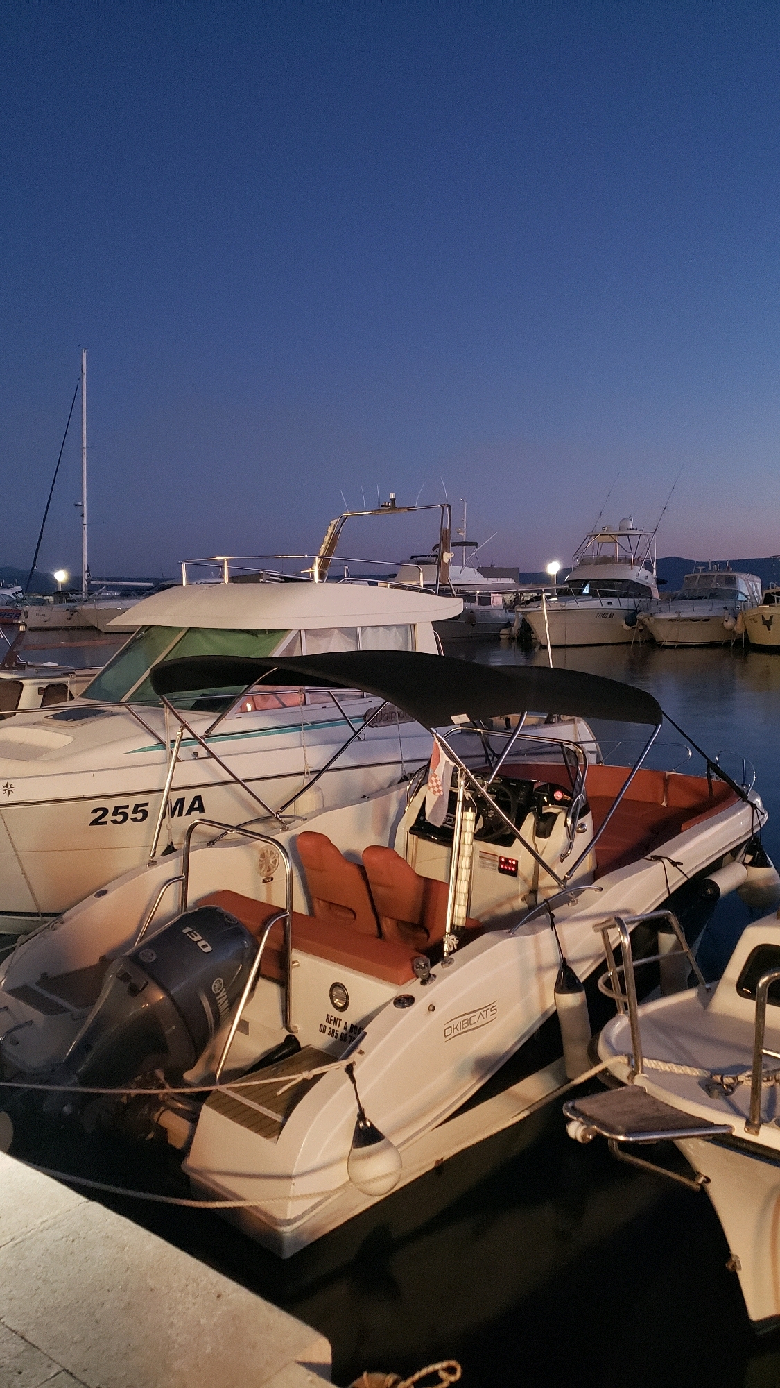 This is a vertical, eye-level, medium shot of several boats docked at a harbor at dusk in Općina Tučepi, Croatia. The sky is a deep blue with a faint gradient towards lighter hues near the horizon. Several boats are visible, some docked in the foreground and others in the background.
In the immediate foreground, a white inflatable boat with orange seating is prominent. It features a Yamaha outboard motor with "130" visible on it. The boat has a black canopy providing shade over the seating area, where a steering wheel and control panel are visible. A Croatian flag is also attached to the boat.
Behind this inflatable boat, a larger white cabin cruiser is partially visible. It has the registration "255 MA" on its side and a green tinted windshield. Several other boats of various sizes and types are docked in the harbor behind these two, some with lights on, suggesting they are preparing for the evening or have just returned. The water is dark and calm, reflecting the lights from the boats and the twilight sky. The scene suggests a peaceful evening at a marina, with the boats secured for the night.