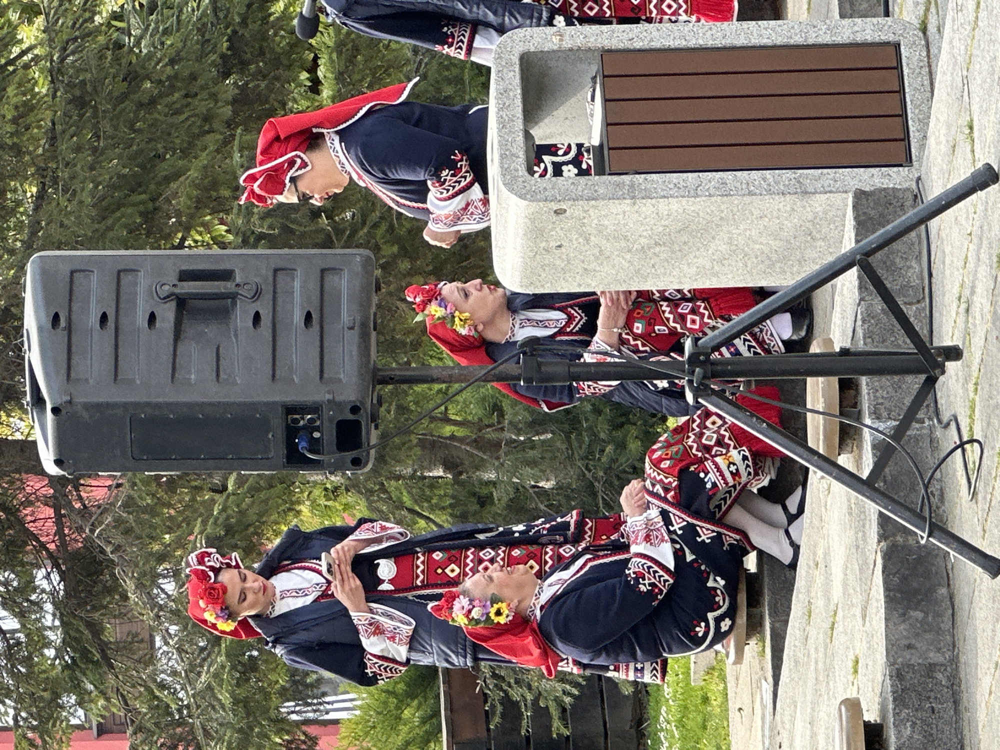 Four women are wearing traditional Bulgarian clothing standing in what appears to be a park or plaza. A woman is standing on a raised stone platform that has a section made of wood. She is wearing a blue outfit with red and white patterns along with a red head scarf. She is looking towards another woman wearing a similar outfit standing below her. To the left, another women is seen wearing similar clothing. In the foreground another woman is seen wearing similar clothing with a flower wreath on her head. A black speaker sits next to the women in the center of the image. The location is likely somewhere in the city of Plovdiv, Bulgaria.