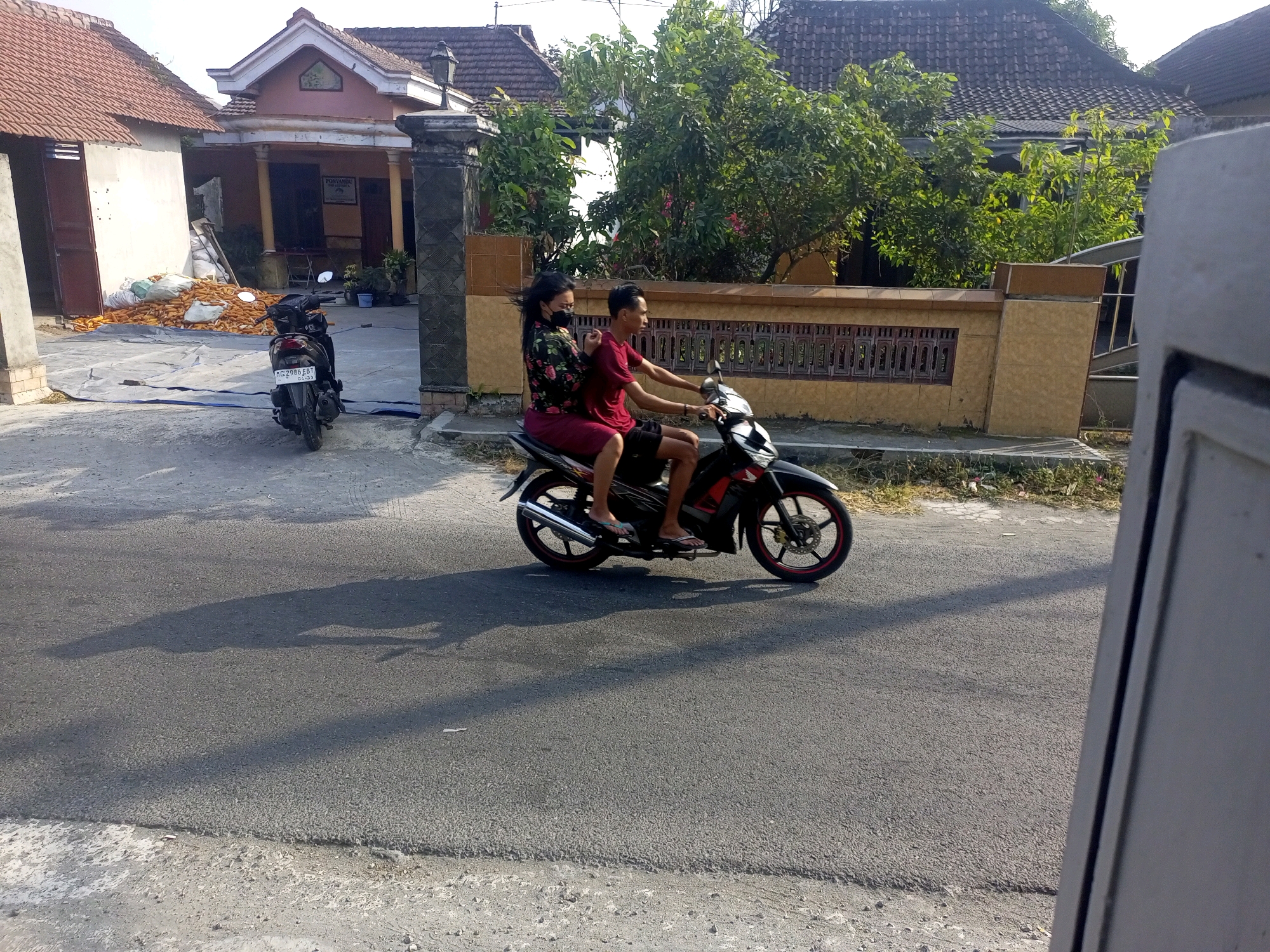 A medium shot shows two people riding a motorcycle on a paved road in Ngasem, Indonesia. The man driving wears a red t-shirt and black shorts. The woman passenger wears a floral print shirt and a red skirt, and a black mask. They are riding past a house with a tiled roof and a decorative wall. In the foreground, a pile of corn is visible next to a building. The lighting suggests it is daytime, possibly late afternoon, given the long shadows. The environment appears to be a rural or semi-urban area.