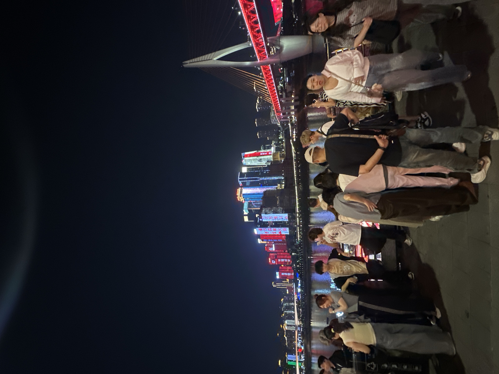 The photo depicts a nighttime scene in Yuzhong District, China, featuring a group of people standing on a waterfront promenade. In the background, illuminated skyscrapers and a bridge with red accents define the cityscape. The lighting is predominantly dark, with artificial lights creating a vibrant contrast against the night sky. People appear to be interacting and observing the urban scenery.