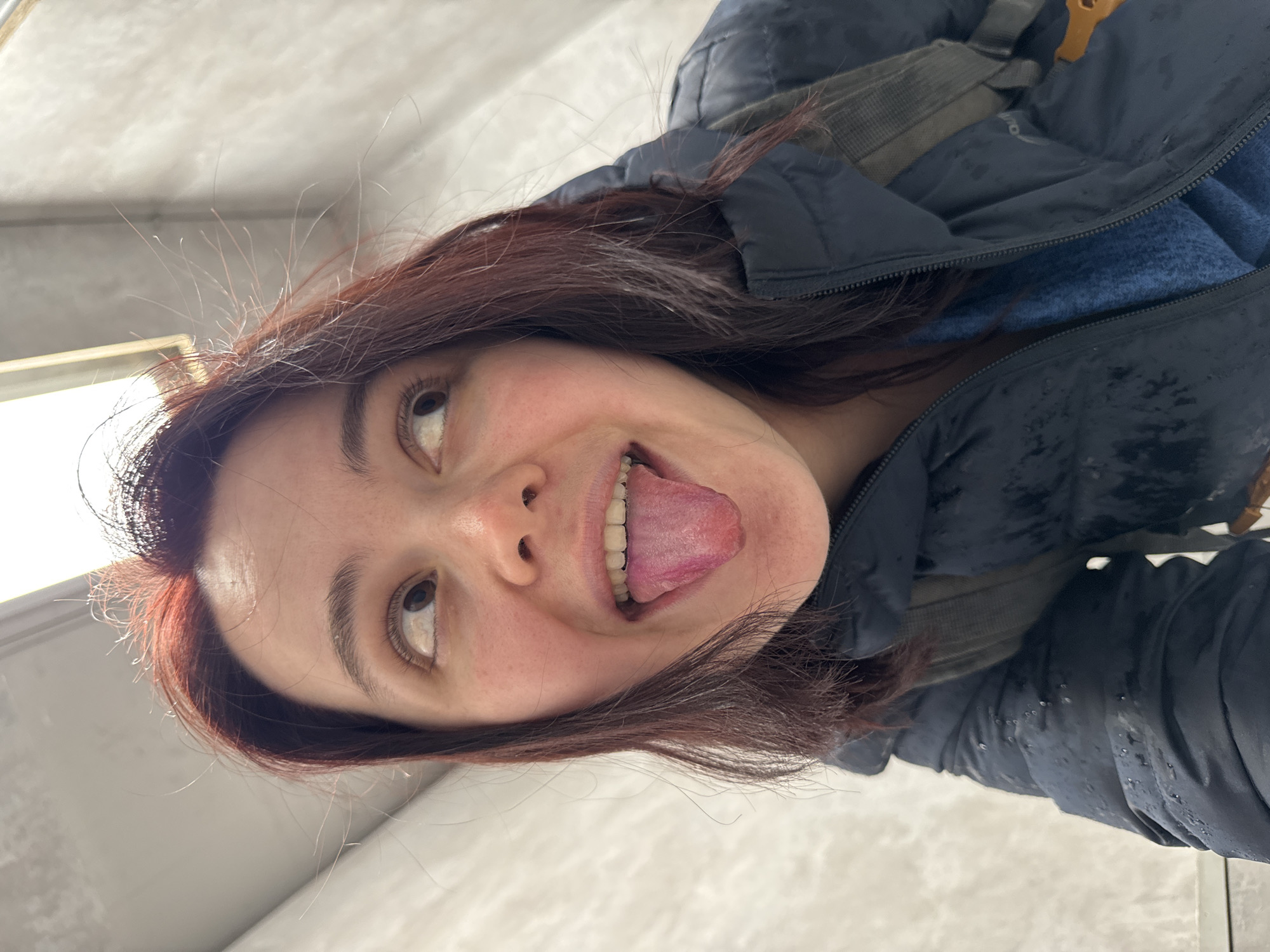 A woman with auburn hair, wearing a dark jacket and a blue top, is sticking her tongue out towards the camera. The image is taken from a low angle, looking up at the woman. The background appears to be a white or light-colored wall and ceiling. There is no evidence in the image to suggest the location is in Izhevsk, Russia.