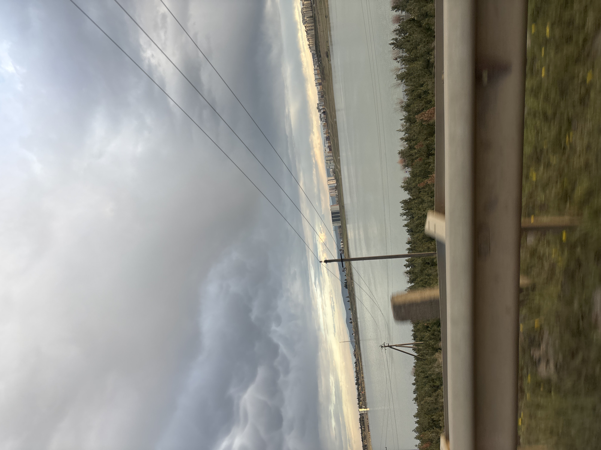 The photograph presents a landscape possibly viewed from a moving vehicle. The sky is overcast with thick, gray clouds dominating the upper half of the frame. Electrical power lines stretch across the sky. Below, a body of water, seemingly a lake or reservoir, reflects the sky's dull light. A line of trees borders the water's edge, suggesting a boundary between the lake and the land. In the distance, a cityscape is discernible, with numerous buildings lining the horizon. The lower right portion of the image features a metal guardrail and a glimpse of roadside vegetation.
Based on the landscape features, specifically the long lake bordered by a city in the distance, this location may be somewhere around Tbilisi, Georgia. It could be near the Tbilisi Sea reservoir.