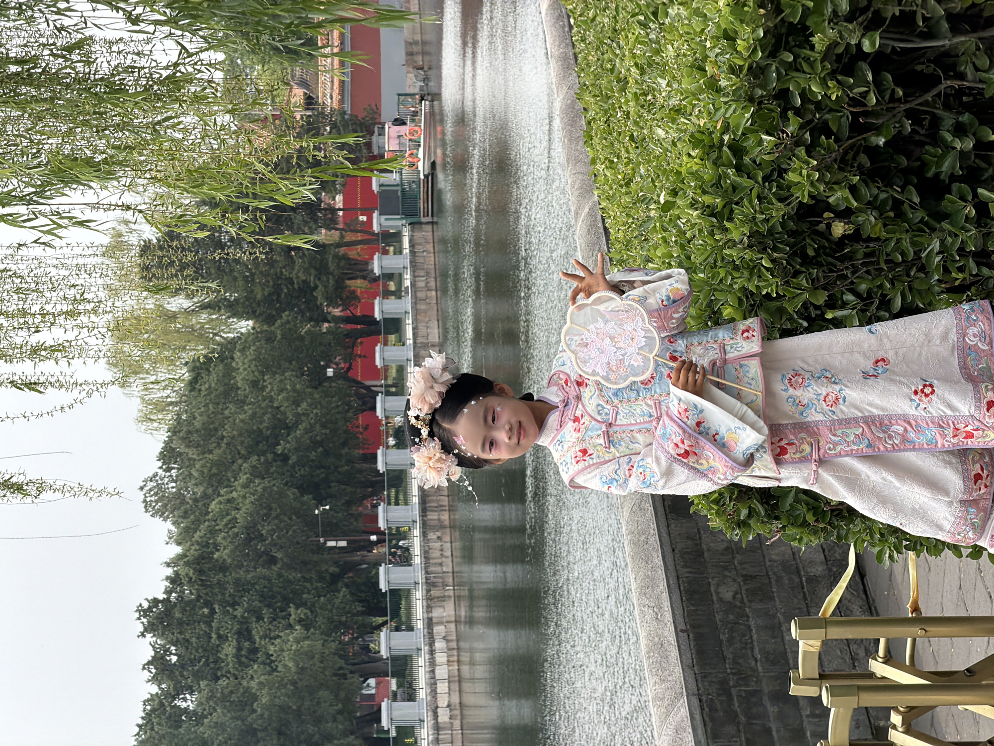 A young girl stands in a traditional Chinese dress alongside a waterway, likely a canal or moat, in the Dongcheng District of China. She is holding a fan and has floral ornaments in her hair. Lush greenery flanks the waterway, with trees and bushes visible. In the background, buildings with traditional Chinese architectural features are present. The location appears to be a park or historical site.