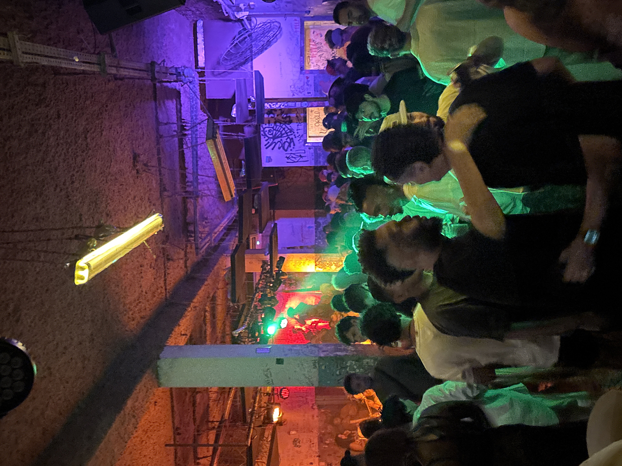 The image depicts a crowded indoor space in what appears to be a nightclub or concert venue. The location is likely in São Paulo, Brazil, based on the metadata. A diverse group of people are gathered, some facing the viewer and others towards a central point, possibly a stage or performance area. Colored lighting, predominantly purple, green, and red, illuminates the scene. The venue's architecture is visibly rough, with exposed brick and support structures. Graffiti and posters adorn the walls. Lighting fixtures hang from the ceiling.
