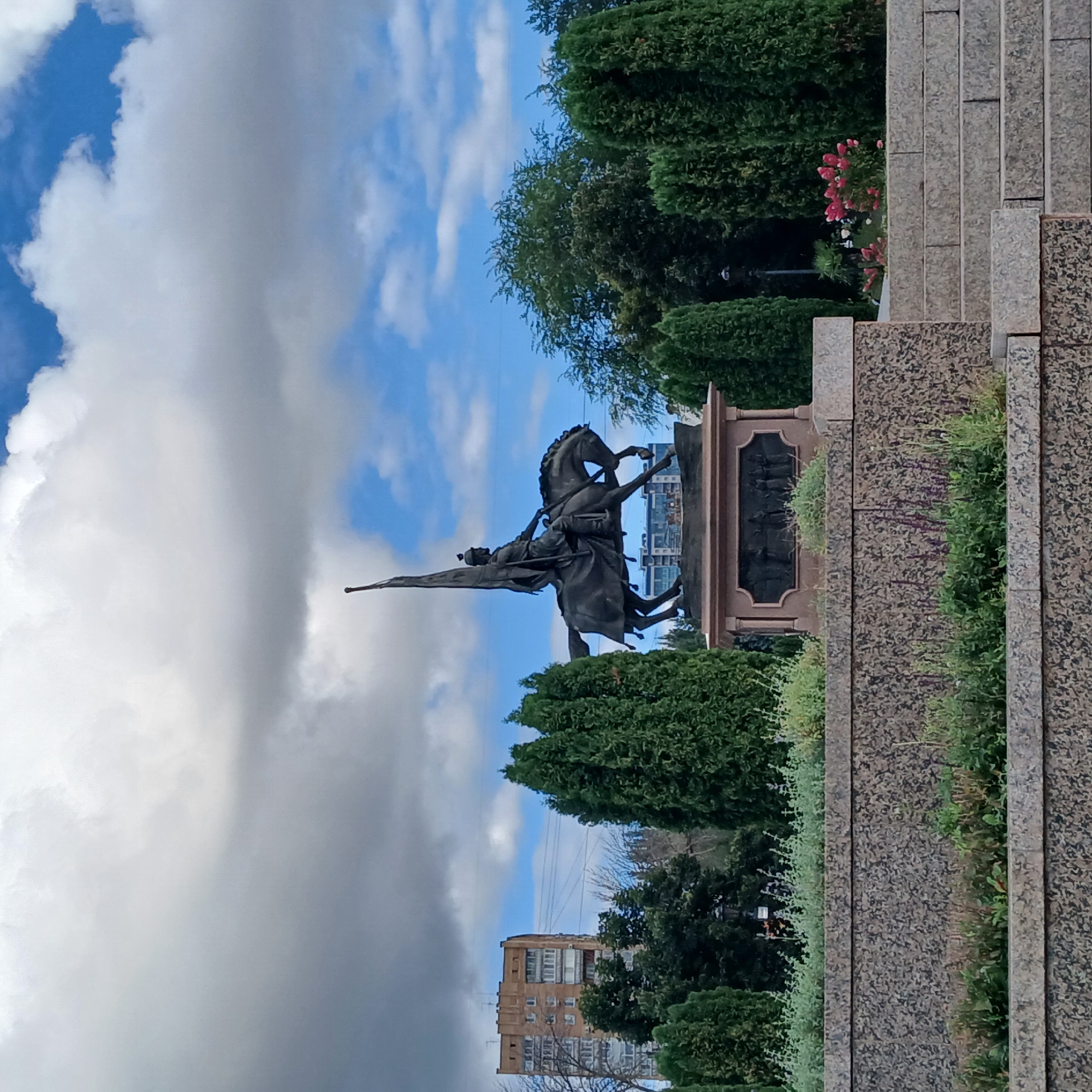 An equestrian statue is centered in the frame, depicting a figure on horseback holding a flag aloft. The statue is made of dark bronze and rests on a stone pedestal which features a bas-relief panel. The scene is set outdoors in what appears to be a park or public square. In the background, several tall, evergreen trees flank the statue, and a multi-story brick building is visible to the left. The sky above is partly cloudy, with patches of blue sky showing through. In the foreground, there is a terraced stone structure with low-lying green plants and some flowering bushes. The image is taken during daylight hours.
This is a photograph of the monument to the Samara Infantry Regiment located in Samara, Russia.