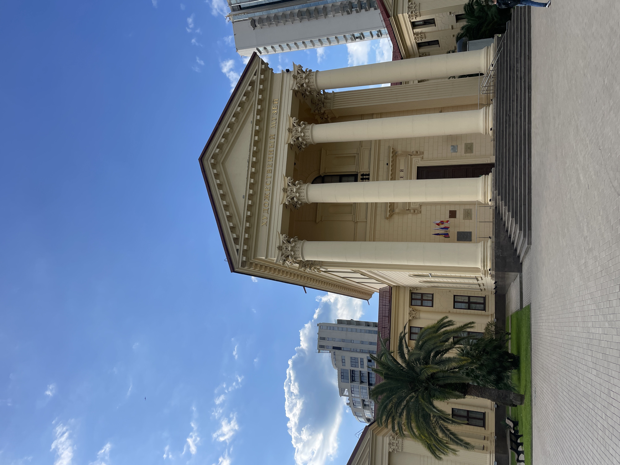The image captures the Sochi Art Museum in Russia. The museum's facade is cream-colored, featuring large white columns and a triangular pediment with text in Russian. A set of stairs leads to the entrance. Flags and plaques are near the base of the columns. A tall modern building is visible in the background. In the foreground, a palm tree is next to the museum's wall. The sky is bright blue with scattered clouds. The camera angle is tilted upward, and a person's legs and feet are visible in the upper right corner of the photo.