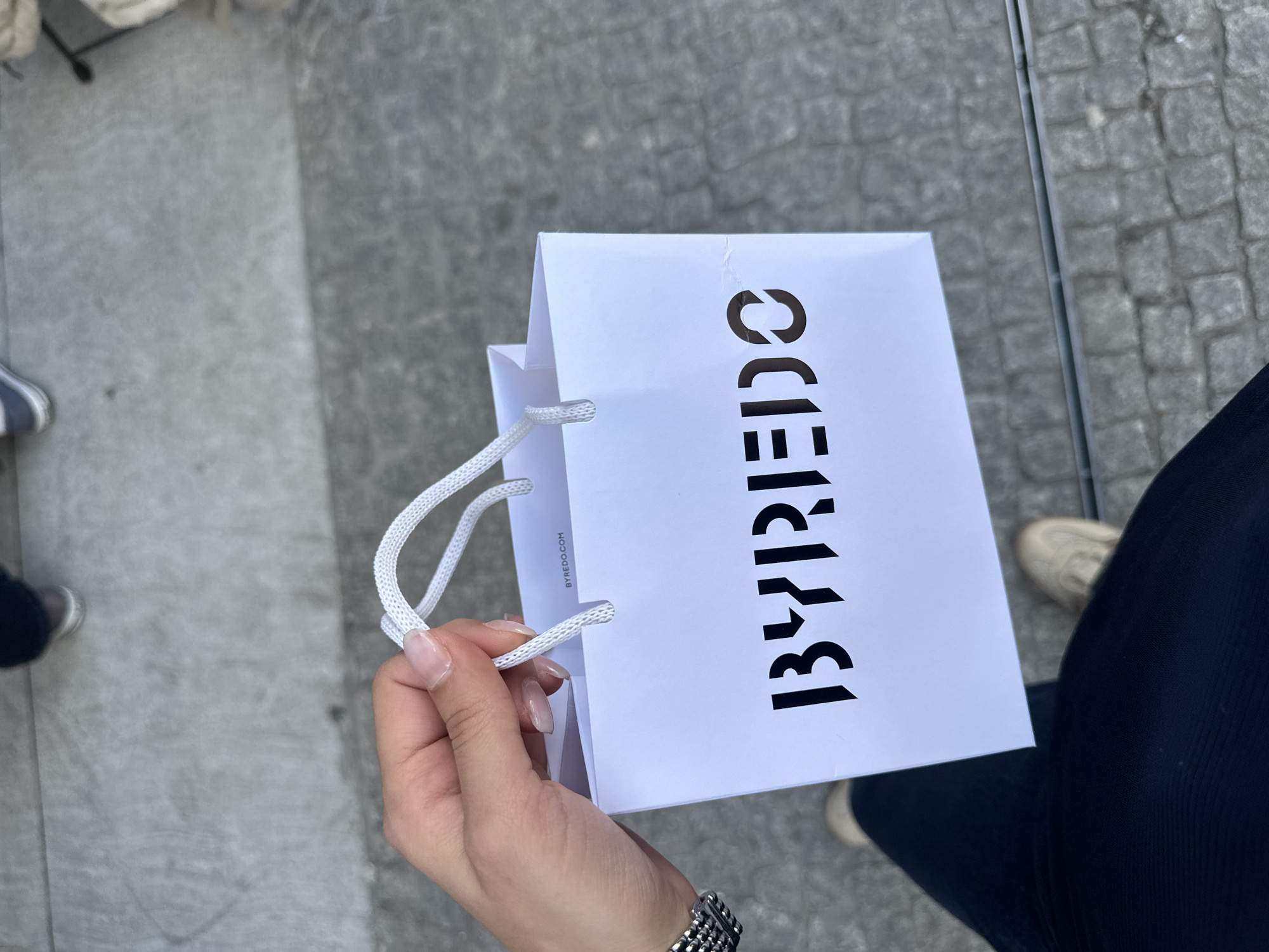 Here's a description of the image:
The image shows a person holding a white Byredo shopping bag, with the brand name printed vertically in black. The bag is held in the person's right hand, which has a silver watch on the wrist. The person is wearing dark blue pants and has tan shoes on their feet. The ground appears to be paved with gray brick or stone. The location is an urban environment, possibly Milan, Italy, given the mention of the city.