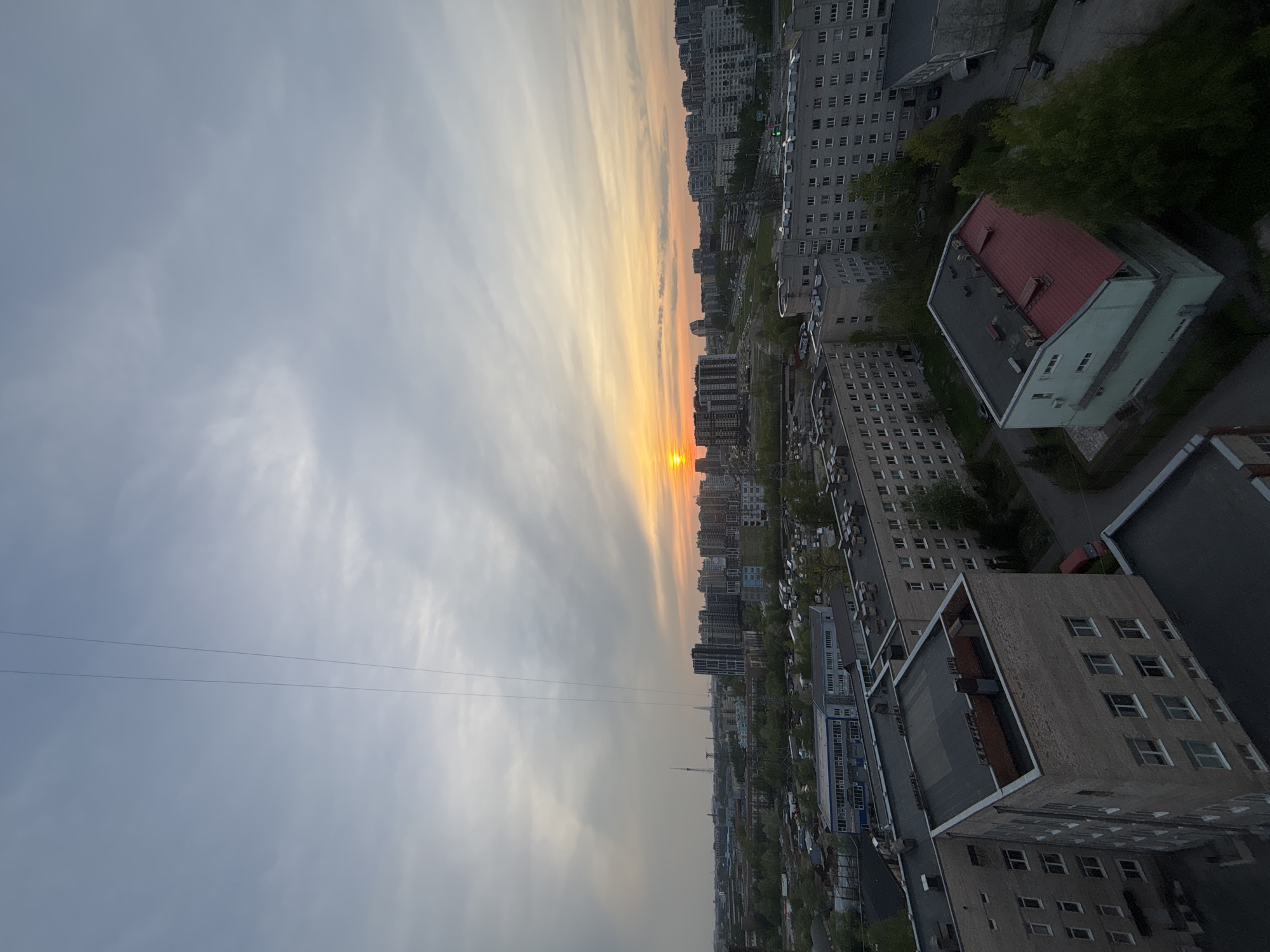 The image shows an urban landscape at sunset. The sky is a blend of gray and orange hues. Below the horizon, there are multiple buildings of varying sizes and architectural styles, indicating a cityscape. There is a line crossing the sky, possibly a wire. The buildings are densely packed together and there appear to be trees interspersed among them.