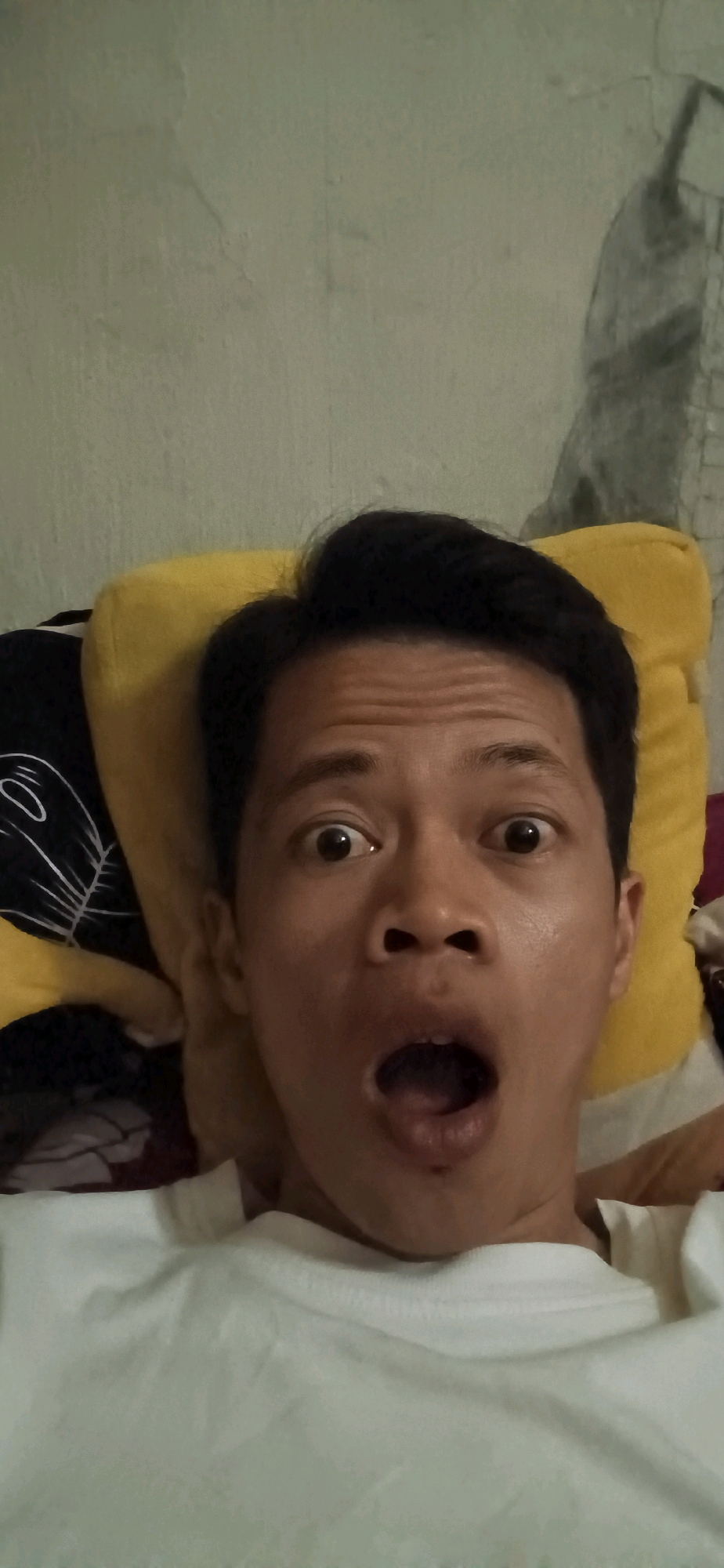 A man is taking a selfie from a low angle, with his mouth wide open in a surprised expression. He is lying down, propped up by a yellow pillow. A portion of a white t-shirt is visible at the bottom of the frame. The background appears to be a wall, possibly in a residential setting in Wonokerso, Indonesia. The image is taken in a dimly lit environment.
