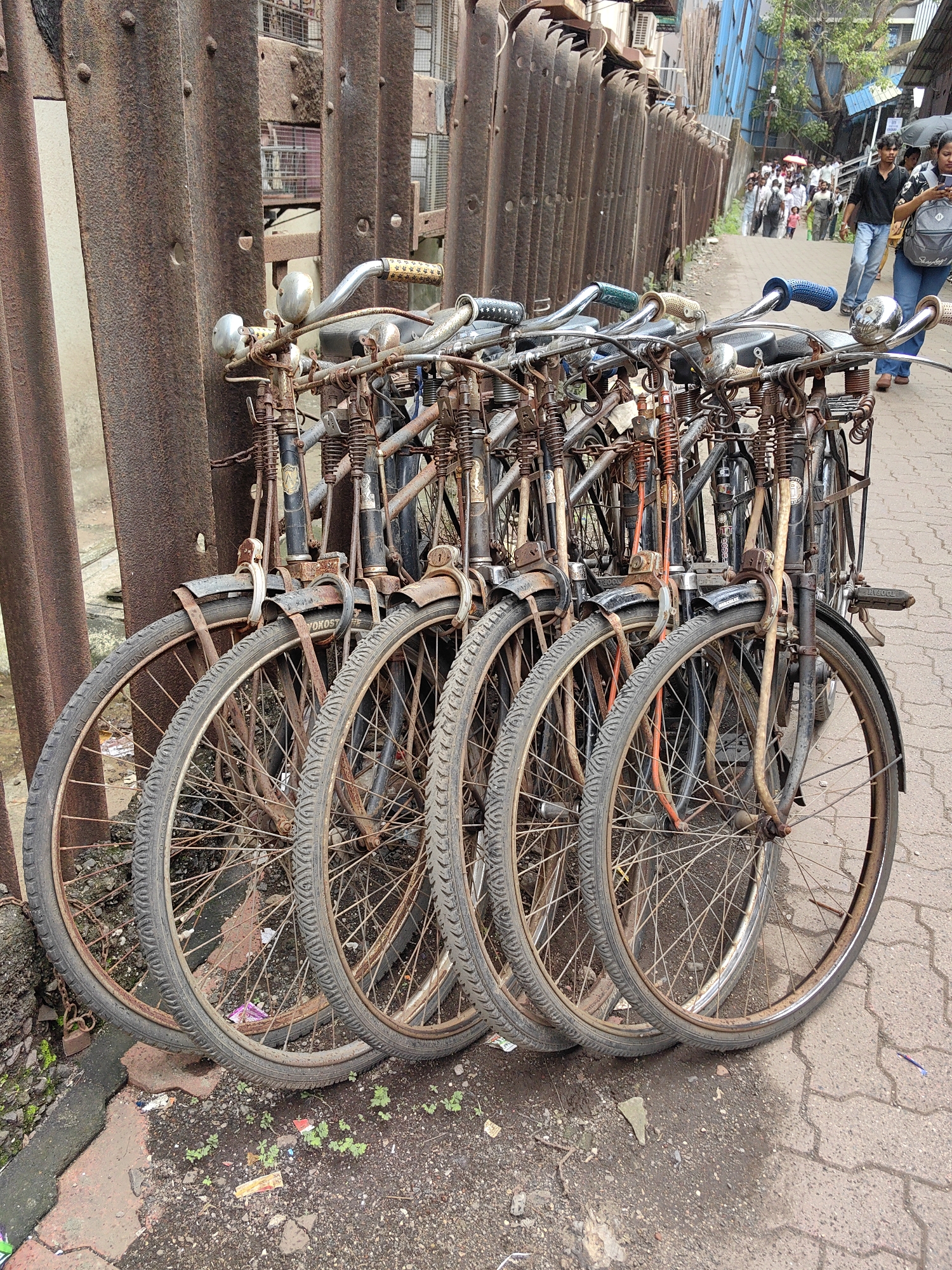In Mumbai, India, several vintage bicycles are lined up and leaning against a rusty, corrugated metal fence. The bicycles are predominantly black and have chrome handlebars, some with colorful grips. They appear to be old and well-used, with visible rust on their frames and spokes. The foreground shows the wheels of the bicycles, with worn tires and spokes. The ground is paved with interlocking bricks and dirt, with some scattered debris like wrappers and small plants. In the background, a street scene unfolds with people walking and a building with blue siding. The overall impression is a gritty, urban environment in Mumbai, showcasing a collection of bicycles parked along a weathered fence.