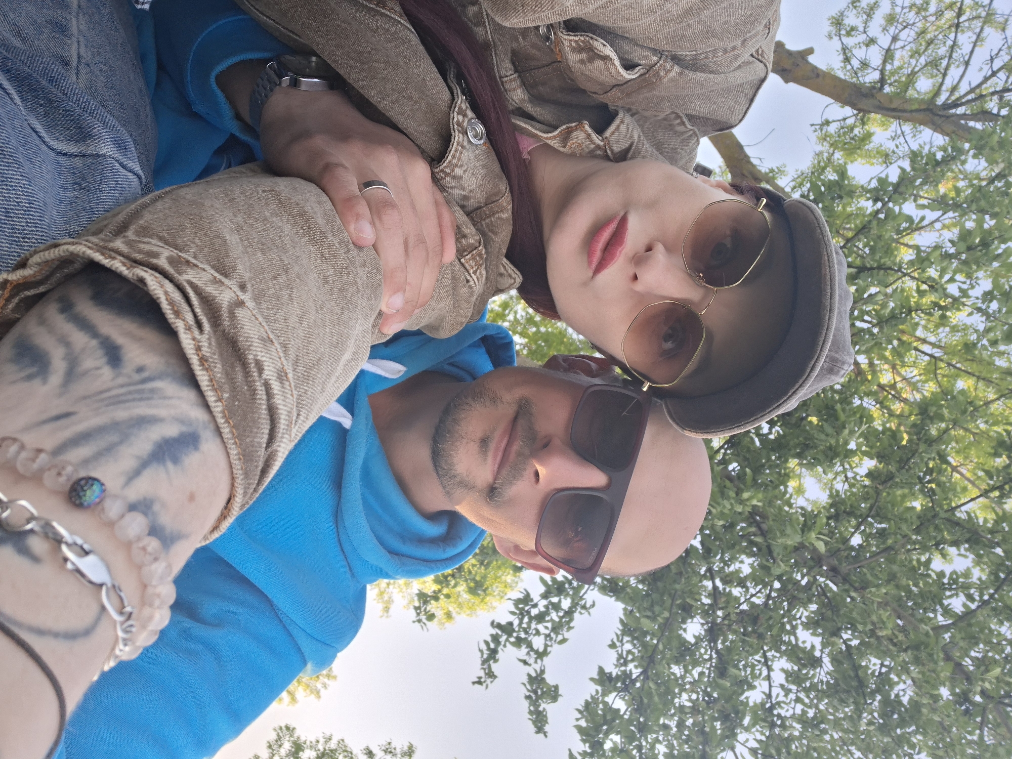 The image is a low-angle selfie taken outdoors in the city of Забалоцкі сельскі Савет, Belarus. Two people are in the frame. A person in a light brown jacket and a hat is at the top of the frame, and a person in a blue hoodie is at the bottom. Both individuals are wearing sunglasses. A tree with green leaves is visible in the background. The person in the blue hoodie has a bracelet and a tattoo on their arm. The person in the light brown jacket has a ring on their finger.