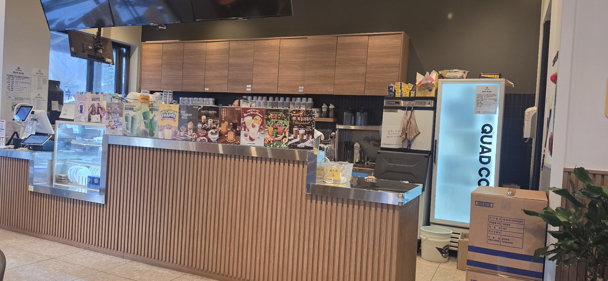 The image shows the interior of a cafe, possibly a franchise, with a modern and clean aesthetic. The cafe features a wooden slatted counter with a stainless steel top. Behind the counter, there is a refrigerated display case with drinks and pastries, and an ice machine. Various beverage posters are displayed on the counter, showcasing popular drink options. A large, illuminated refrigerator with the text "QUAD CO" is visible on the right side of the image. In the background, there are wooden cabinets and shelves stocked with supplies. Several cardboard boxes are stacked near the refrigerator, suggesting a recent delivery or preparation for shipment. The overall atmosphere is bright and inviting, typical of a contemporary coffee shop. The presence of Korean text on posters and labels indicates the location is in South Korea, potentially Cheongju-si, as per the prompt. No people are clearly visible, except for a partial view of someone behind the counter.