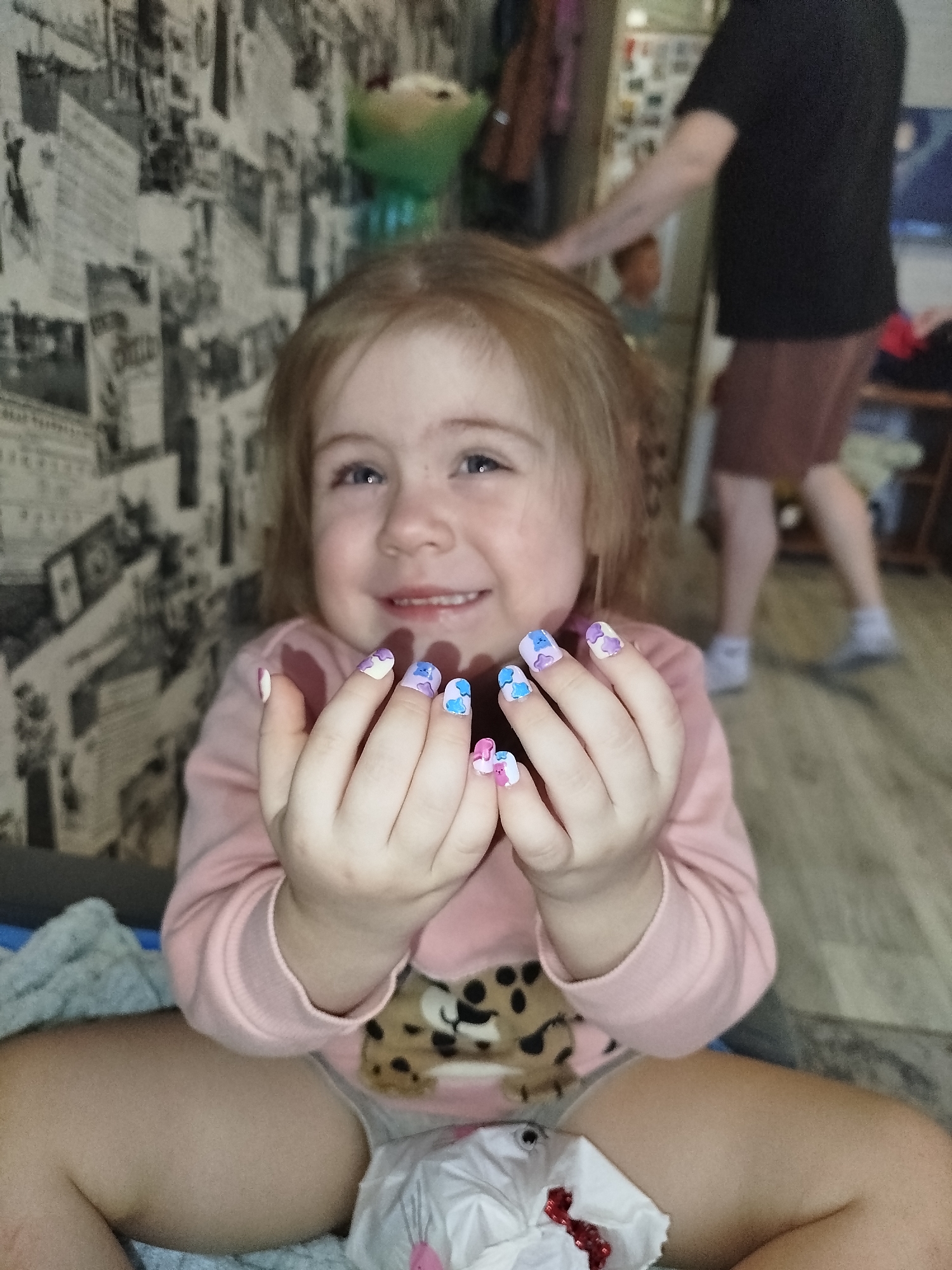 A young child is sitting indoors, displaying decorated fingernails. The child has fair skin, light brown hair, and is wearing a pink long-sleeved shirt with a cartoon animal design. The fingernails are adorned with a pattern of blue, purple, and pink. In the background, an adult wearing a black shirt and maroon shorts is visible, walking on the floor. A toy can be seen on a shelf behind the child. The wallpaper is decorated with a black and white newspaper-style print. A used tissue with red remnants is on the child's lap. The location is Chaikovsky, Russia.