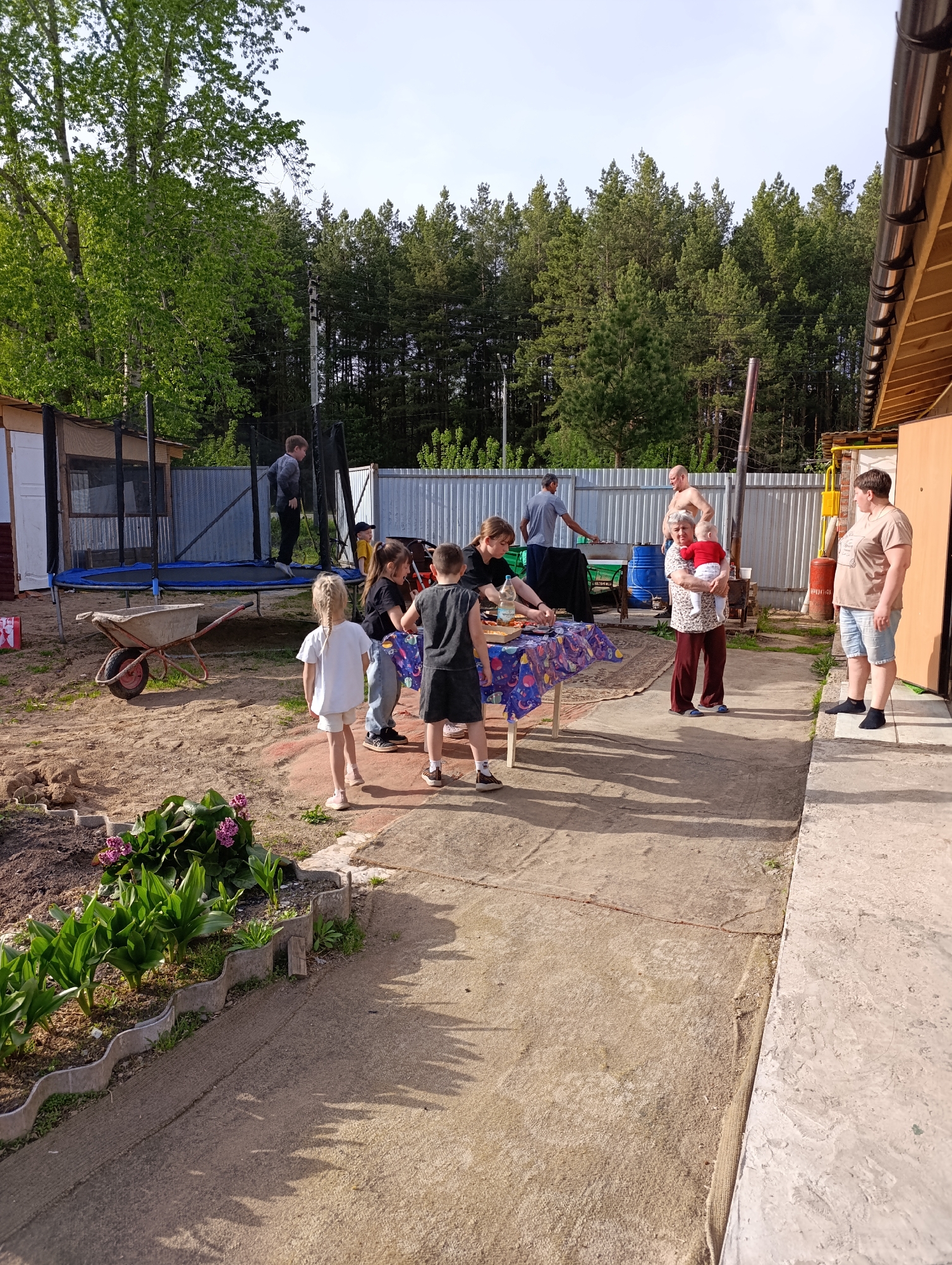 The image shows an outdoor gathering in what appears to be a backyard in Chaikovsky, Russia. In the foreground, there is a garden bed with flowers. A concrete patio extends from the bottom of the frame towards the center, where a table is covered with a patterned tablecloth. Around the table, several children are gathered. Behind them, an older woman is holding a baby. To the right, a person is standing near the side of a building. In the background, other individuals are near a grill and a fence. A trampoline is visible on the left side, with a person bouncing on it. A wheelbarrow is also present near the trampoline. The area is surrounded by trees.