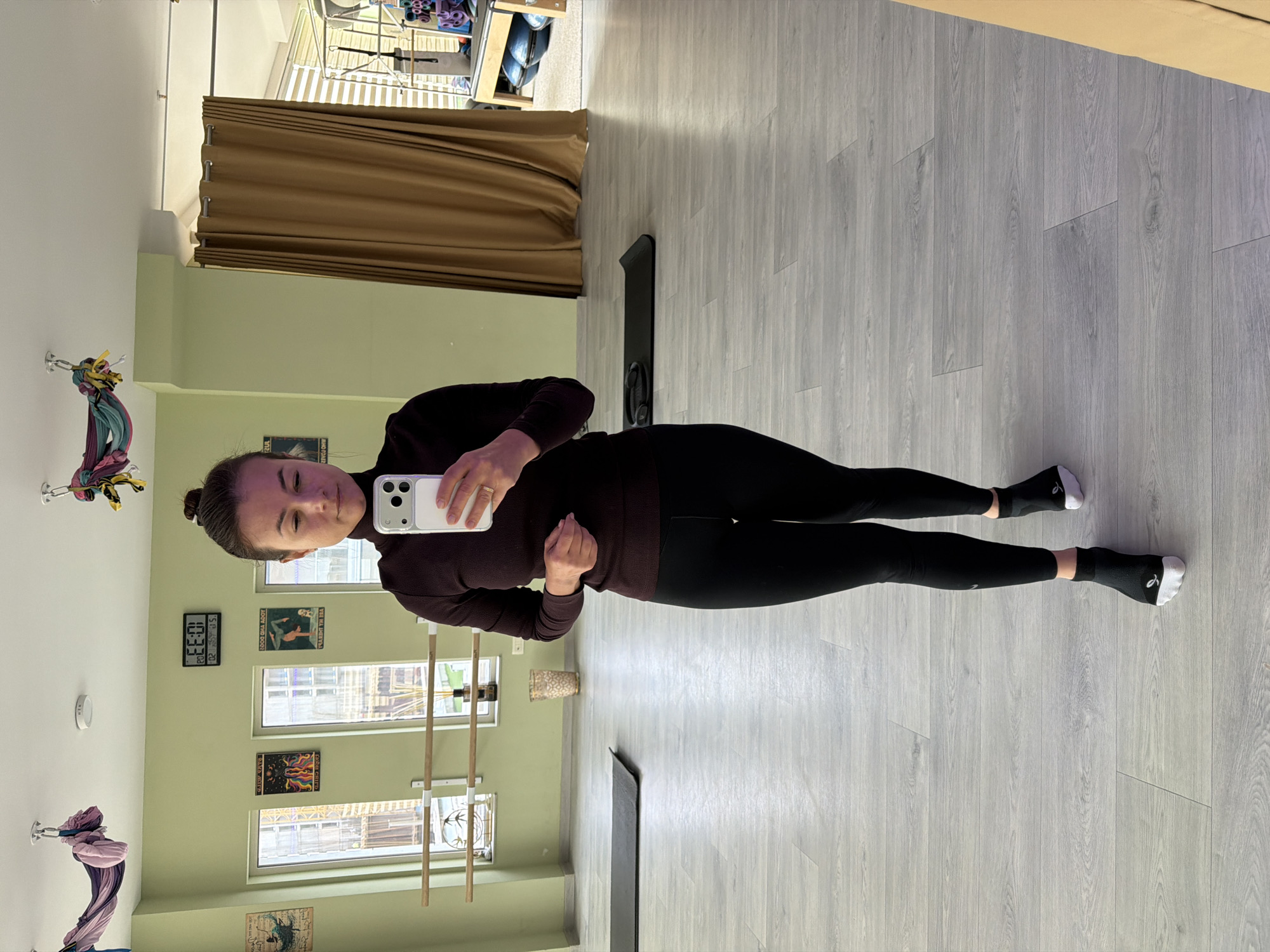 A woman is taking a selfie in a dance studio. She is wearing a dark purple turtleneck sweater and black leggings with black socks that have white heels and toes. The studio has light green walls and a light gray wood-look floor. In the background, there are windows with a ballet barre in front of them and posters on the wall. There is also a dark brown curtain, possibly dividing the studio space. In the reflection of a mirror, ceiling mounted dance/yoga silks are visible. A digital clock on the wall shows the time as 10:33. Without further information, the location in Budva, Montenegro cannot be confirmed.
