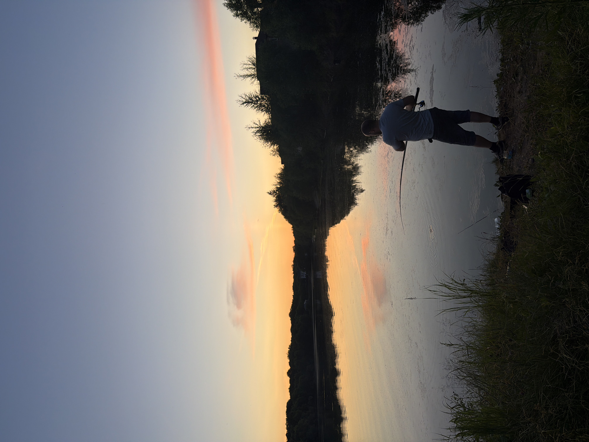 This image depicts a tranquil scene at dusk by a body of water, with a lone fisherman casting a line. The sky exhibits a gradient of soft blues and oranges, with hints of pink from scattered clouds, indicative of sunset. The water's surface mirrors the sky's colors, creating a serene reflection. Along the far bank, a line of trees forms a dark silhouette against the illuminated sky, with a few buildings partially visible through the foliage, suggesting a rural or semi-rural setting. In the foreground, tall grasses and dense vegetation line the bank. A man, wearing a grey t-shirt and dark shorts, stands facing the water, holding a fishing rod in his hands, engaged in the act of fishing. A backpack rests on the ground beside him. The overall atmosphere is one of peaceful solitude and quiet recreation. The location is identified as Birobidzhan, Russia.