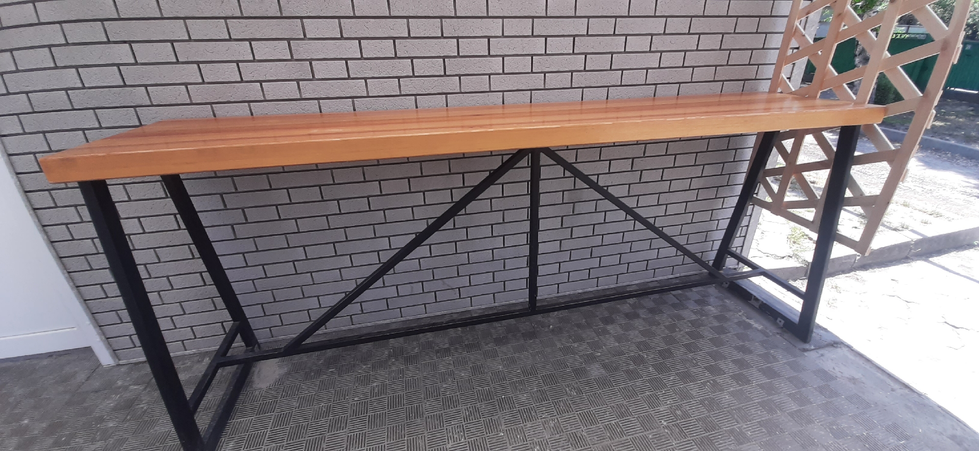 A wooden-topped bar-height table with a black metal frame stands outdoors against a light grey brick wall. The table has a slatted wooden seat attached to the back of its surface, forming a partial backrest. The metal frame is constructed of vertical legs connected by horizontal and diagonal supports. The table is situated on a patterned grey floor. In the background, sunlight illuminates an outdoor area with cracked concrete and some vegetation. The location is Yessentukskaya, Russia.