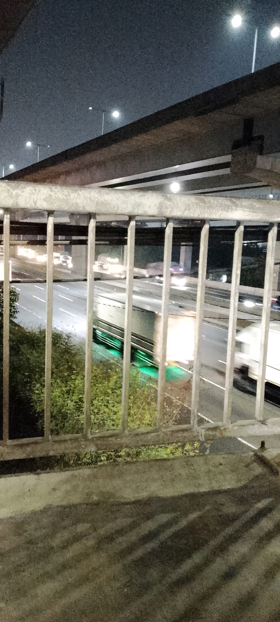The image captures a night scene of a highway in Bekasi, Indonesia, viewed from an elevated walkway with metal railings in the foreground. The highway is illuminated by streetlights, and traffic is visible with streaking headlights and taillights indicating movement. One truck, in particular, has green undercarriage lights. There are trees and foliage on the side of the highway. Above the highway, another elevated structure, possibly a bridge or overpass, is visible against the night sky, which is dotted with streetlights. The overall impression is of a busy urban highway at night.