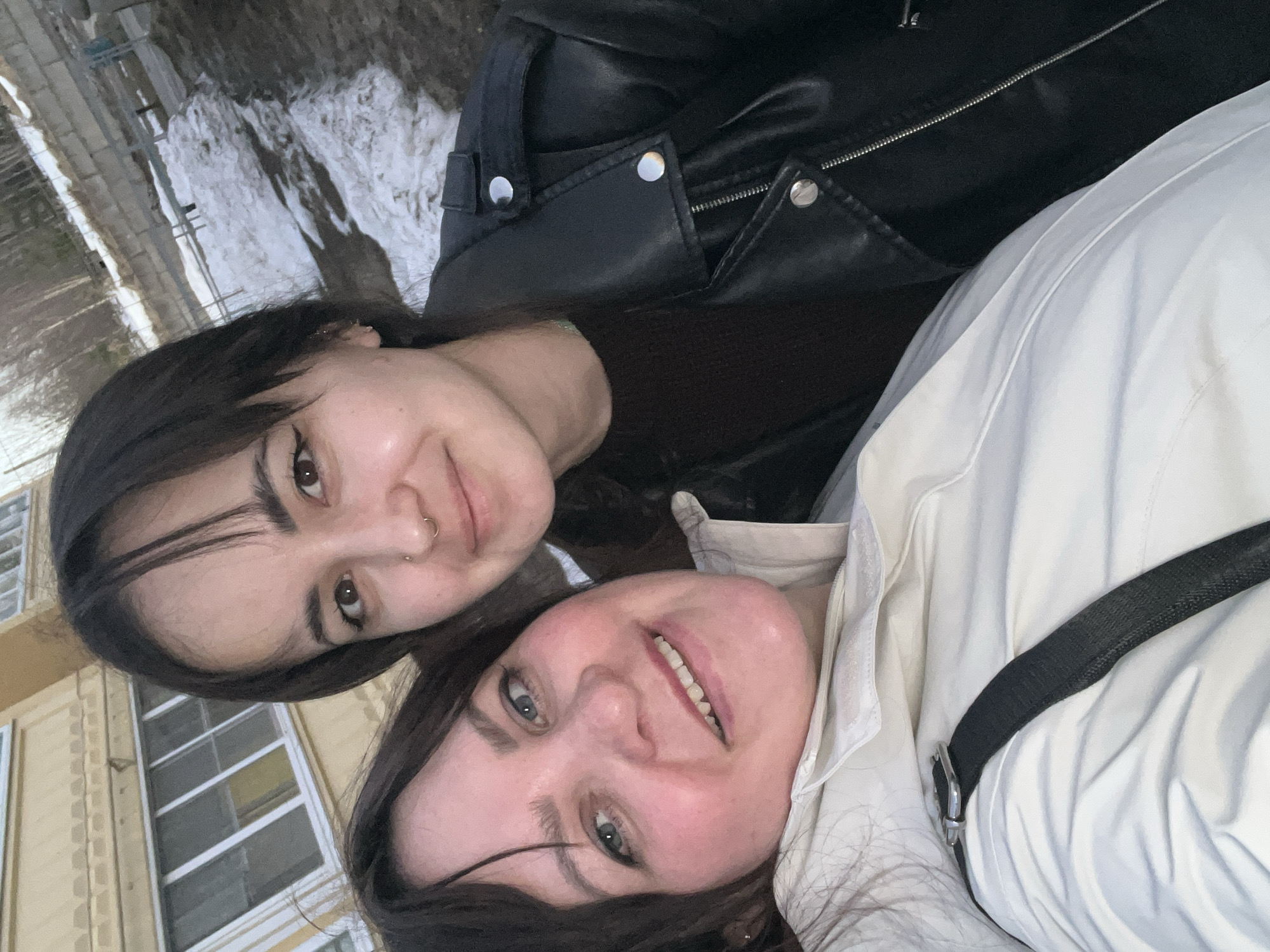 Two women are posing for a selfie outdoors. The woman on the left has dark hair and is wearing a black leather jacket. The woman on the right has dark hair and is wearing a white jacket. The background shows a building with windows and snow on the ground. The city is Noviy, Russia.
