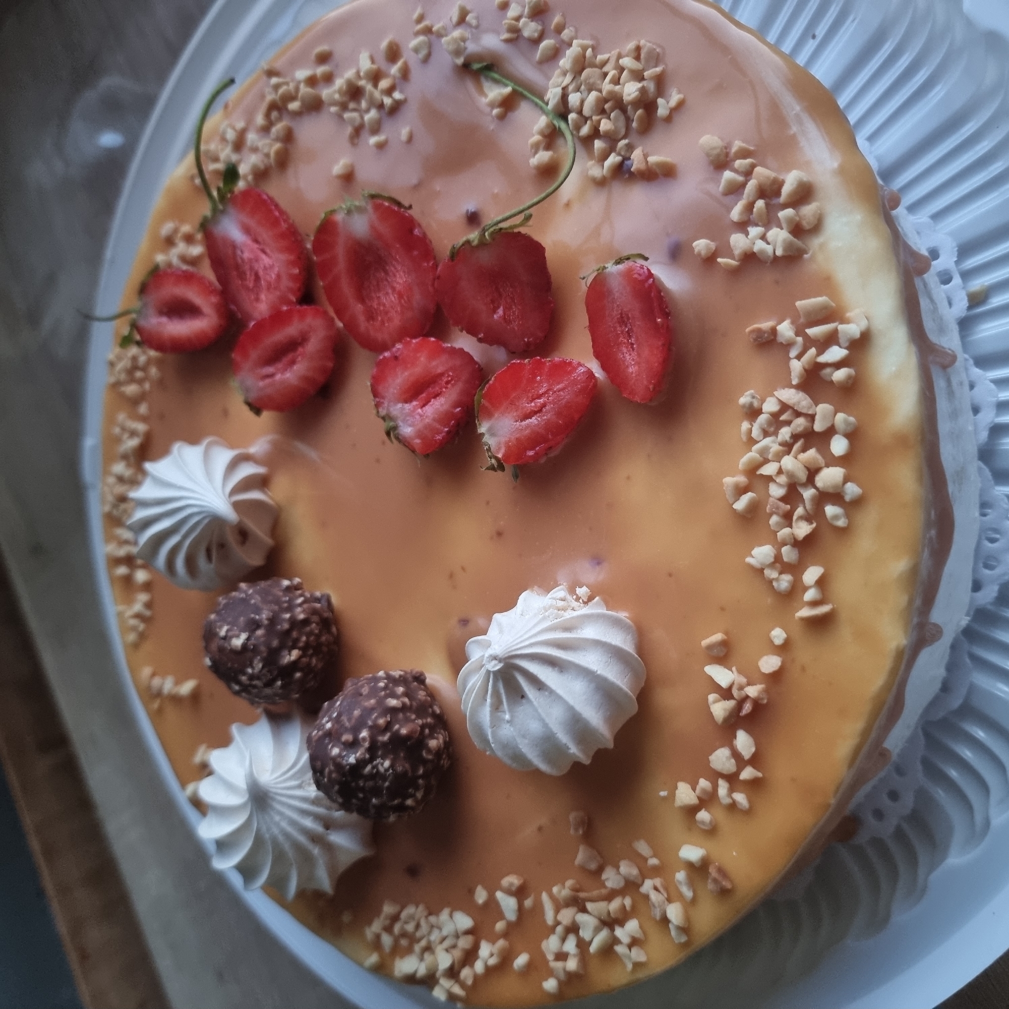 A circular cake with a glossy caramel glaze is presented on a white cake stand. The cake is adorned with several halved strawberries, small white meringue swirls, and chocolate-covered hazelnut candies. Chopped peanuts are sprinkled across the top of the cake. The cake appears to be indoors, possibly on a wooden table, as suggested by the background. The scene is shot from a high angle, focusing on the details of the cake's decoration. There are no people or animals visible in the image, and no specific location in Aktobe, Kazakhstan, can be identified.