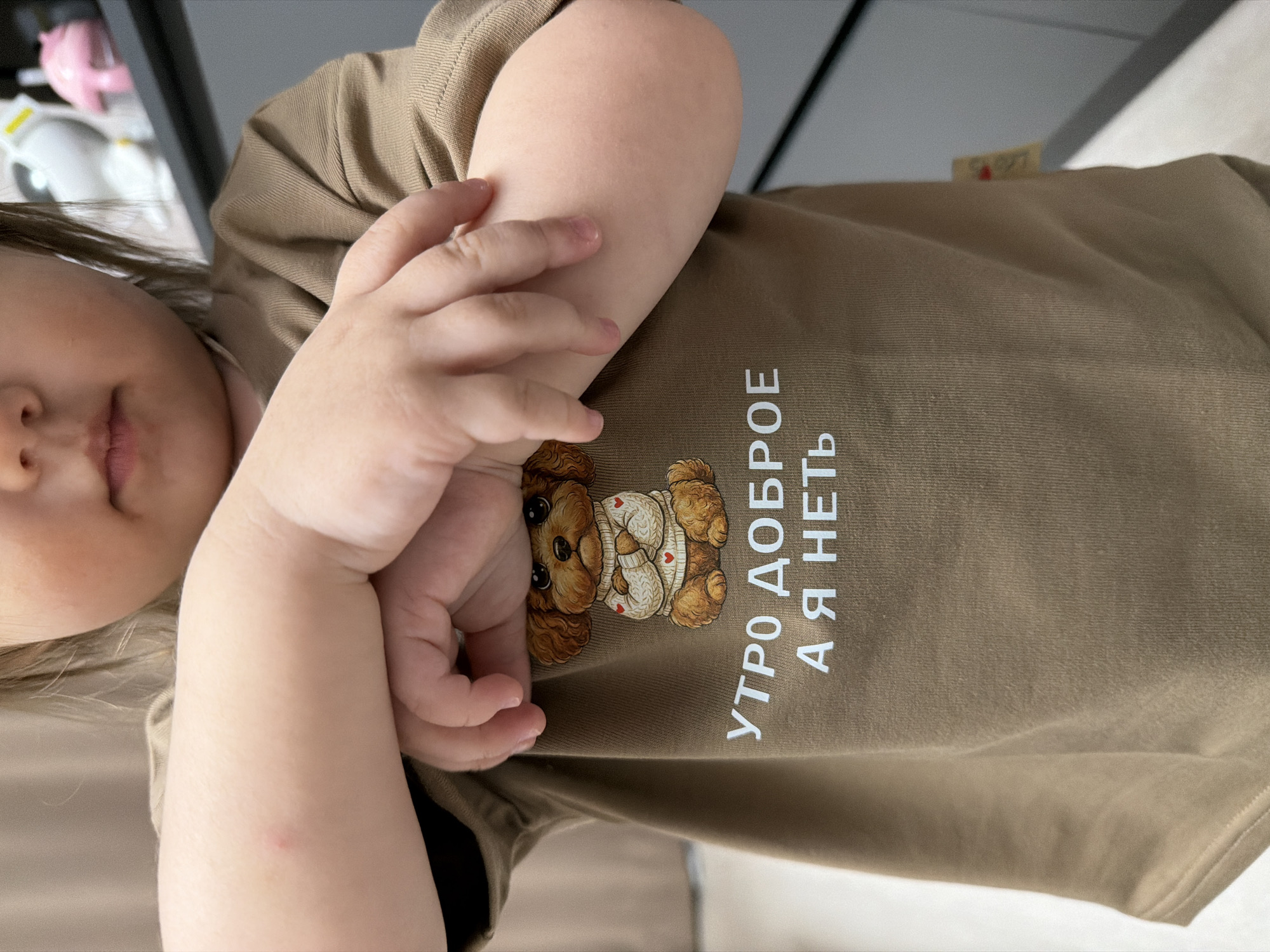 An indoor scene in Reshetnikovo, Russia, depicts a young child from the chest upwards, primarily focusing on their torso and arms. The child's lower face, including the mouth and chin, is visible. They are wearing a light brown or tan long-sleeved shirt featuring a graphic of a brown, fluffy dog, possibly a poodle, wearing a cream sweater adorned with multiple small red hearts. Below the dog, white Cyrillic text reads "УТРО ДОБРОЕ А Я НЕТ," which translates to "Good morning, but I'm not." The child's arms are crossed in front of their chest, with their hands resting on the opposing forearms. A small, distinct red mark is visible on the inner aspect of the child's left forearm. The background consists of a light-colored, out-of-focus wall or curtain on the left and a dark shelf with partially visible white and pink decorative figurines, including a white object with ears and a pink mushroom-shaped object, on the right.