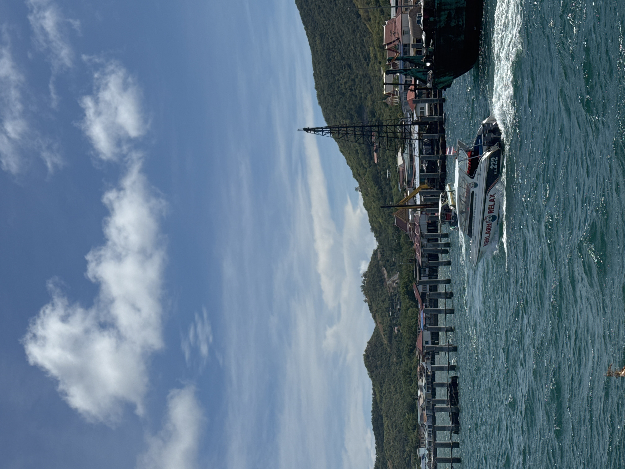The image captures a coastal scene, likely in Pattaya City, Thailand. In the foreground, turquoise water churns around a white and green motorboat with the identifier "KL222". The boat is labeled "KOH LARN RELAX". Adjacent to the water, a pier extends into the sea, and buildings line the coast. Further inland, a green, forested mountain provides a backdrop to the urban development. Above, a blue sky is dotted with white clouds.
