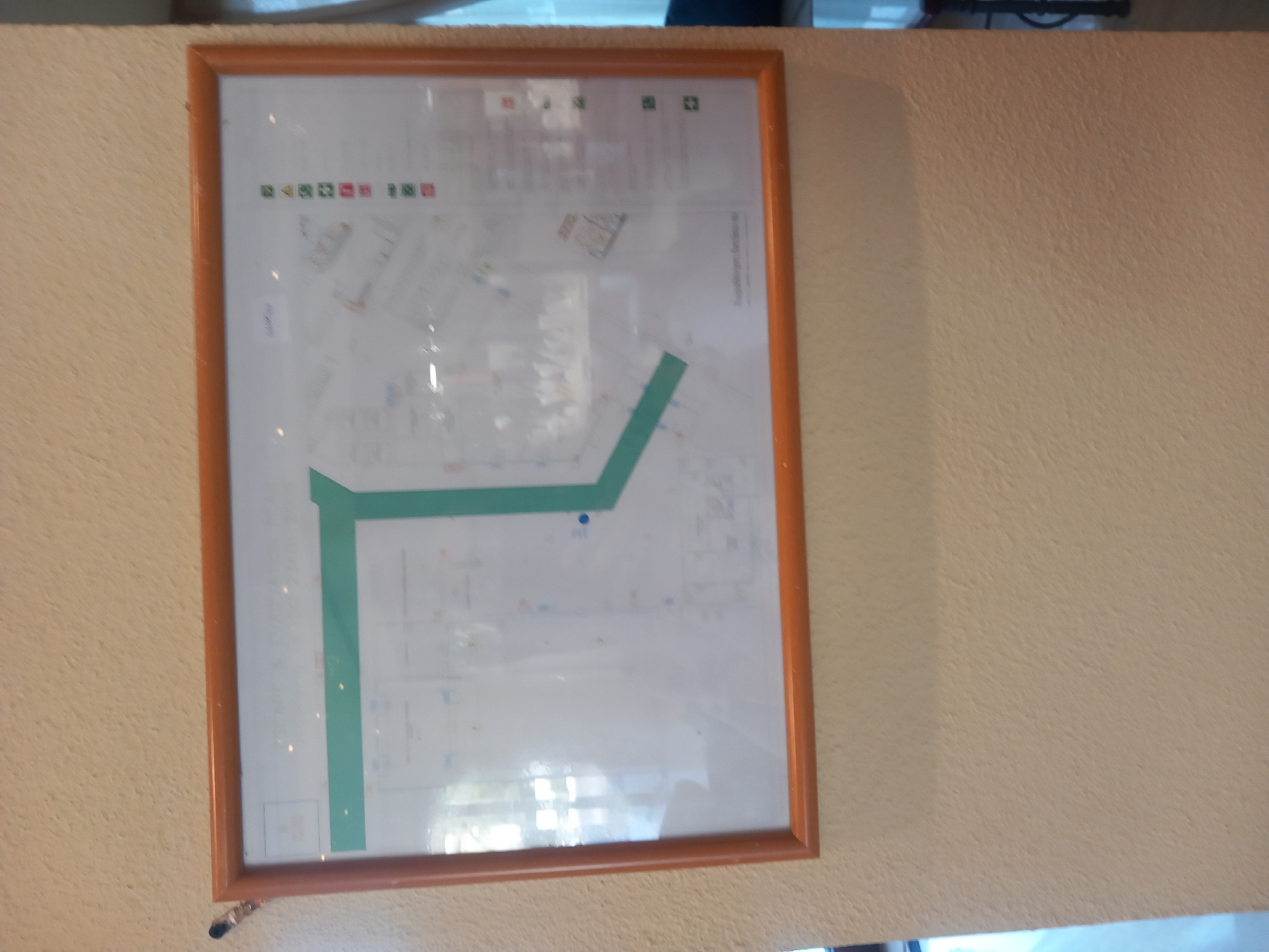 A framed diagram, likely a floor plan or emergency exit map, is mounted on a textured, light beige wall. The diagram features a prominent green pathway, indicating a route. To the right of the diagram, there is a vertical list of small colored symbols, including green squares, a red triangle, and red squares. The diagram is encased in a wooden frame with a warm, reddish-brown hue. The wall has a rough, stucco-like texture. The upper left corner of the frame is slightly disturbed by a black object, possibly a tack or pin, with a small silver component. Light reflections are visible on the surface of the diagram, suggesting it is under glass or a similar protective material. The overall scene appears to be indoors, possibly in a public building or establishment. The location is identified as the city of Kos, Greece.