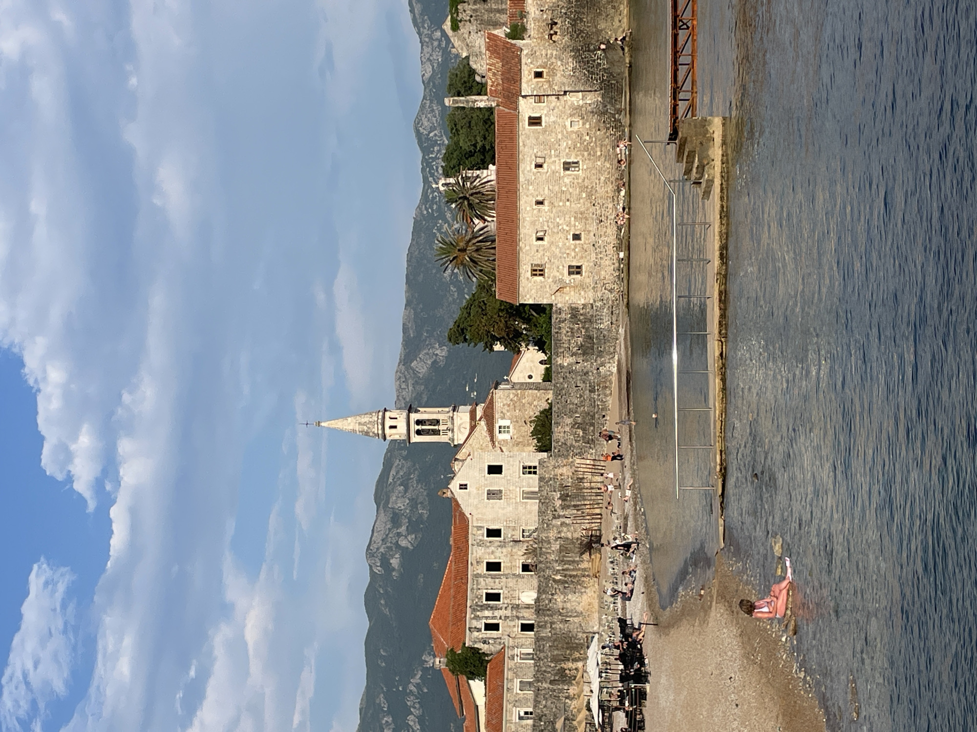 The image depicts the island of Sveti Stefan, a luxury resort in Budva, Montenegro, with buildings constructed of light-colored stone and red-tiled roofs. A tall church spire rises above the buildings, set against a backdrop of mountains. The sky is partly cloudy, with blue patches. The Adriatic Sea borders the island. On the right side of the image, a concrete walkway and a metal ladder lead into the water. Several people are relaxing or swimming at the edge of the water. Some are near the wall, while one person is partially submerged in the sea. At the bottom of the image, there are dark objects that appear to be tables and chairs in front of a building.
