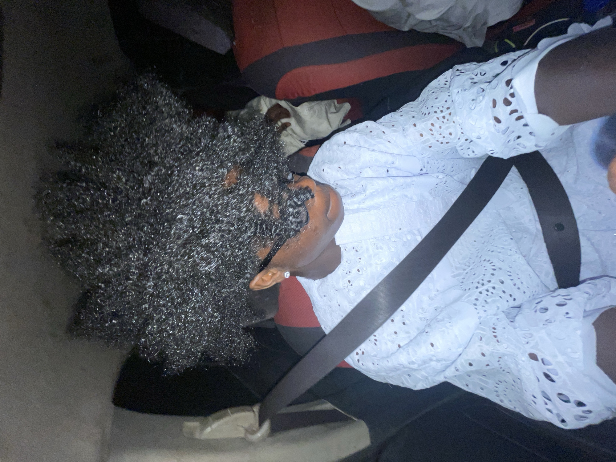 A person is seated in a car, wearing a white, eyelet-patterned shirt, a black seatbelt, and black pants. The person has dark curly hair, and a light-colored earring is visible on the left ear. The car seat is red and black. The photo was taken inside a car, presumably in Igboefon, Nigeria.