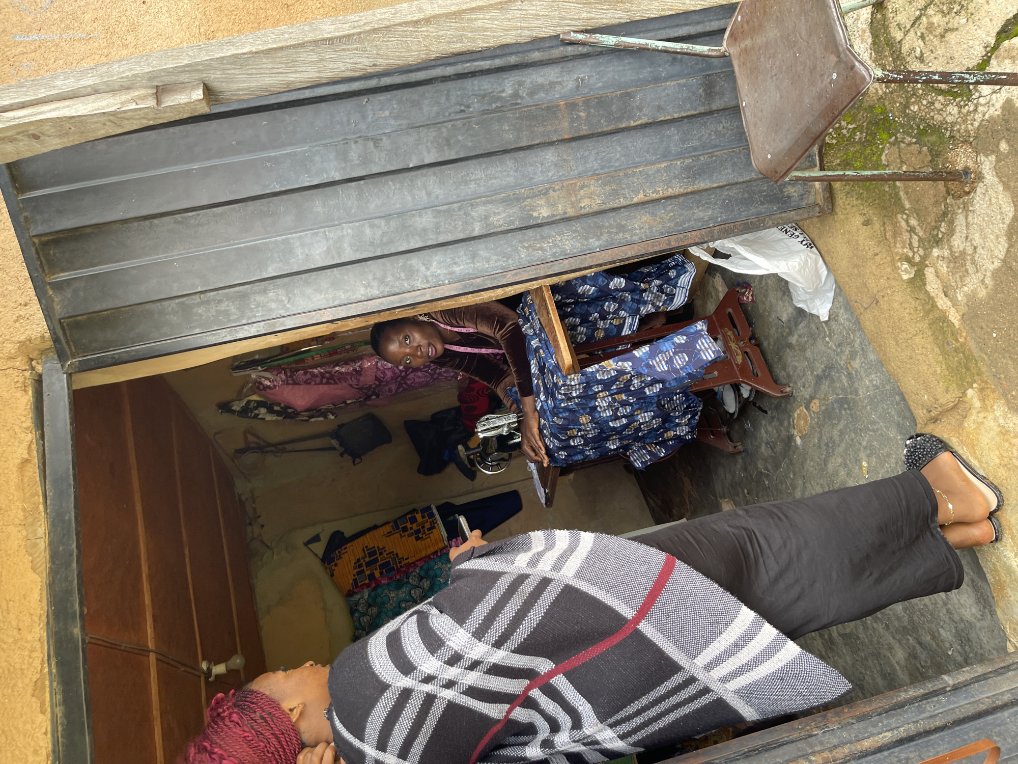 The image depicts an indoor scene, likely a workshop or a tailoring studio, in Rafin Par, Nigeria. Two individuals are present. In the foreground, a person wearing a black skirt and a patterned shawl draped over their shoulders is lying down or reclining. Their head is turned to the side, and their feet are visible, adorned with black, embellished flats and a delicate anklet.
Behind this person, further inside the space, another individual is seated. They are wearing a brown velvet top and a blue and white patterned skirt. This person is looking towards the camera with a slight smile. A pink measuring tape is visible around their neck. They are positioned near a sewing machine, suggesting a tailoring or dressmaking activity.
The space itself appears somewhat cluttered but organized, with stacks of fabric, including the blue and white patterned material, and other textiles visible. A white plastic bag is also present. The walls are rough, and the entrance seems to be a metallic roller shutter door, partially open, revealing the interior. The lighting suggests it is daytime, though no specific time can be determined. The overall mood appears casual and perhaps industrious. No visible text is decipherable.